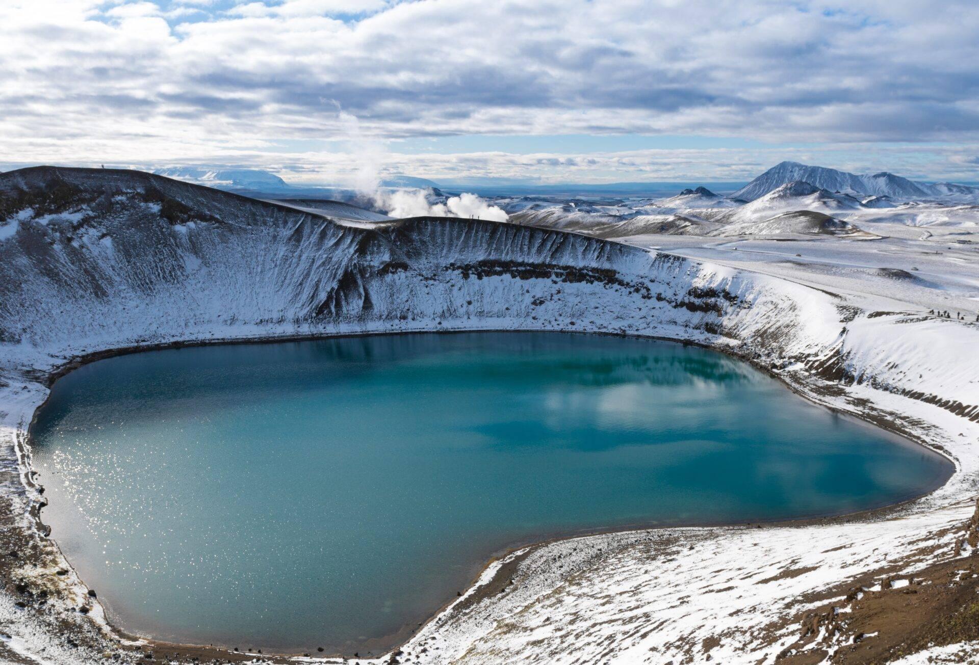 Un cratere vulcanico innevato con un lago turchese al centro, circondato da montagne innevate sotto un cielo nuvoloso