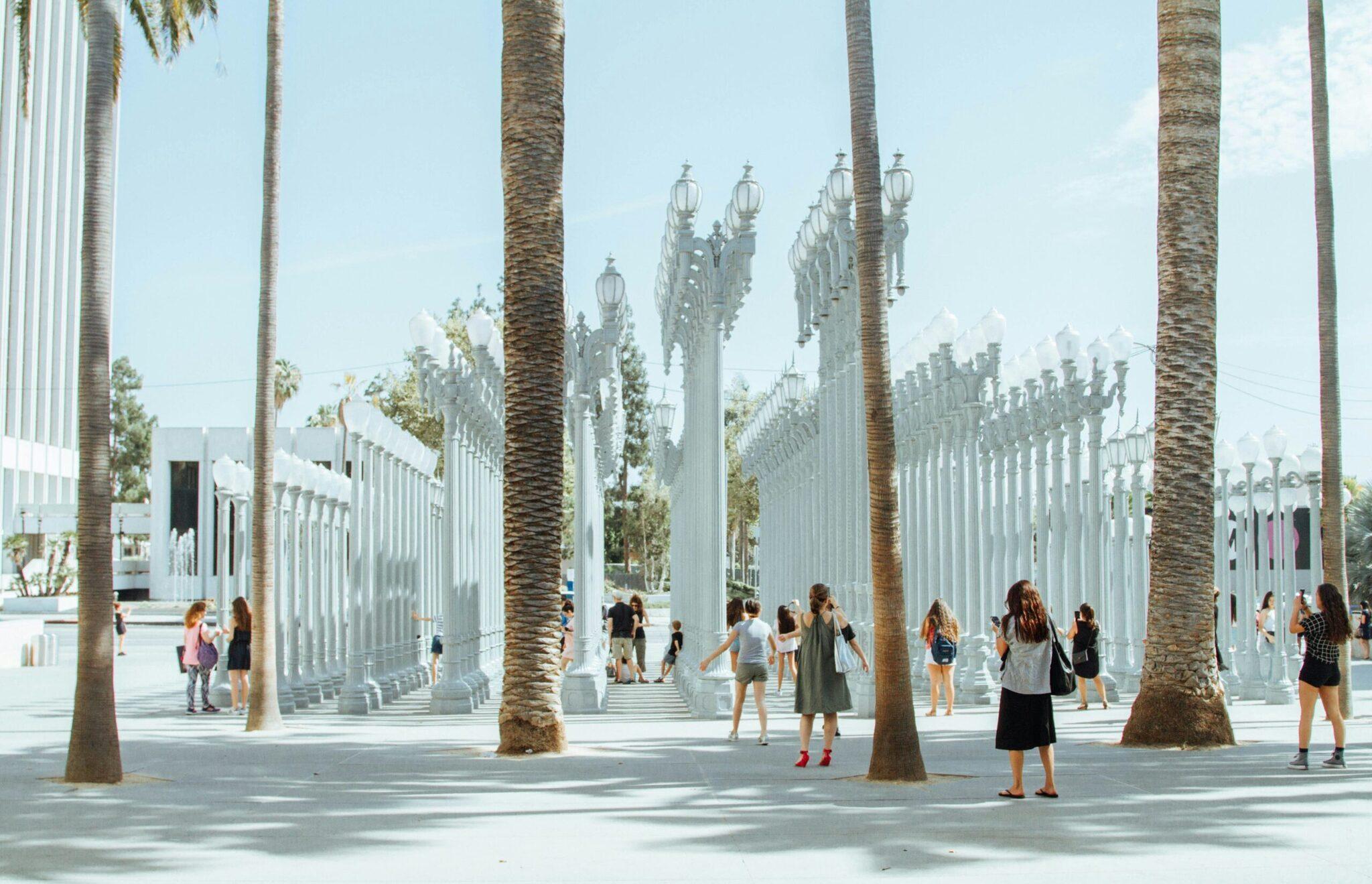 Eine moderne öffentliche Plaza mit weißen Säulen, Palmen und Straßenlaternen, belebt von Besuchern unter einem klaren Himmel