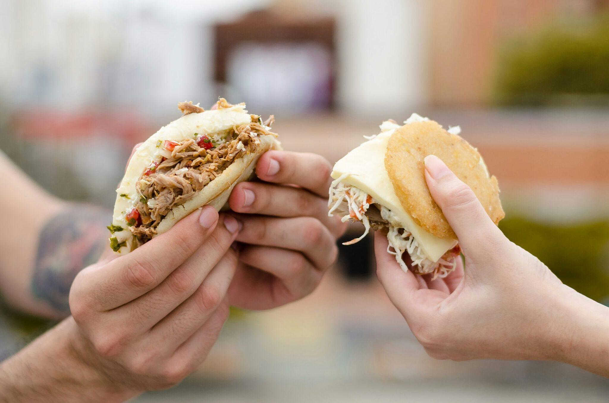 Deux mains tenant des sandwichs garnis de viande effilochée dans une rue pavée en arrière-plan flou