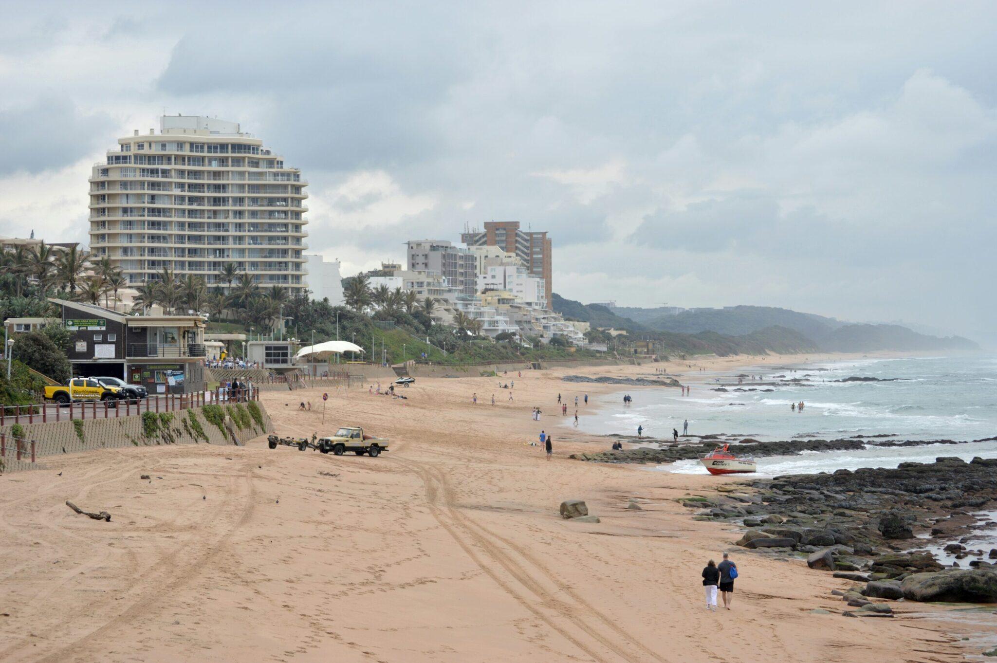 Une plage côtière avec bâtiments résidentiels, rochers noirs, baigneurs et véhicules sur le sable