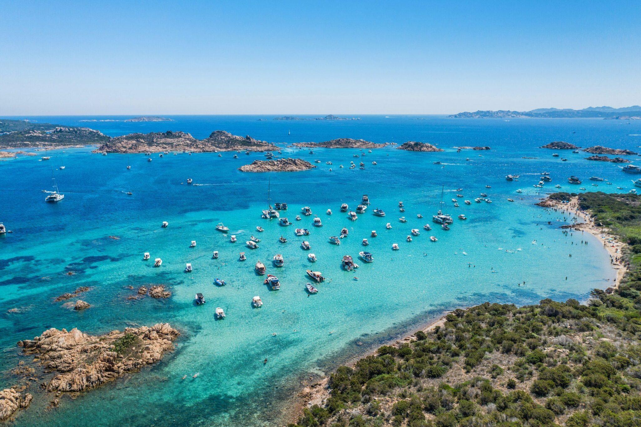 Vue aérienne d’une baie turquoise parsemée de bateaux amarrés, entourée d’îlots rocheux et de côtes méditerranéennes verdoyantes