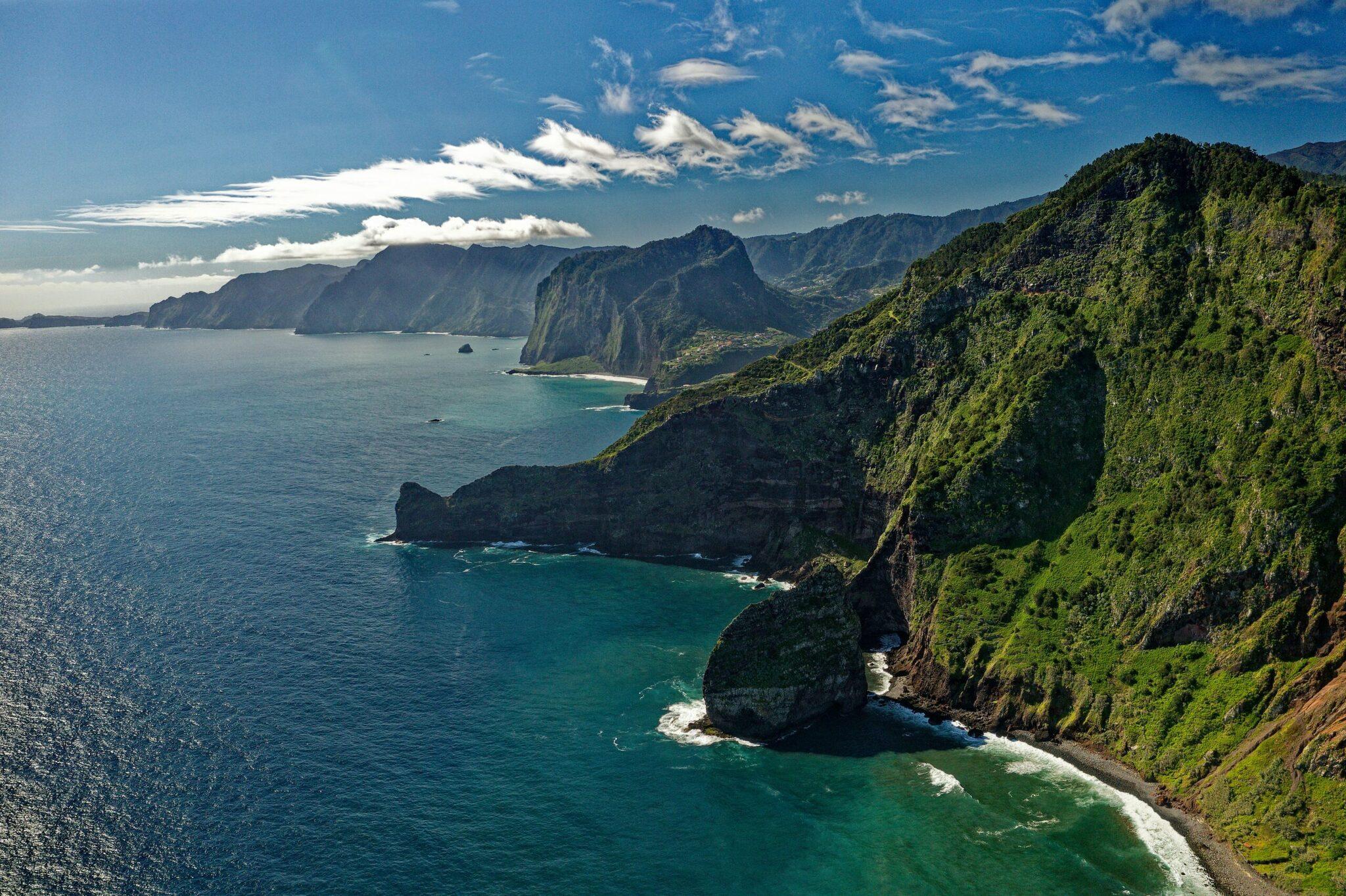 Falaises côtières verdoyantes dominant une mer turquoise avec montagnes volcaniques en arrière-plan sous ciel bleu