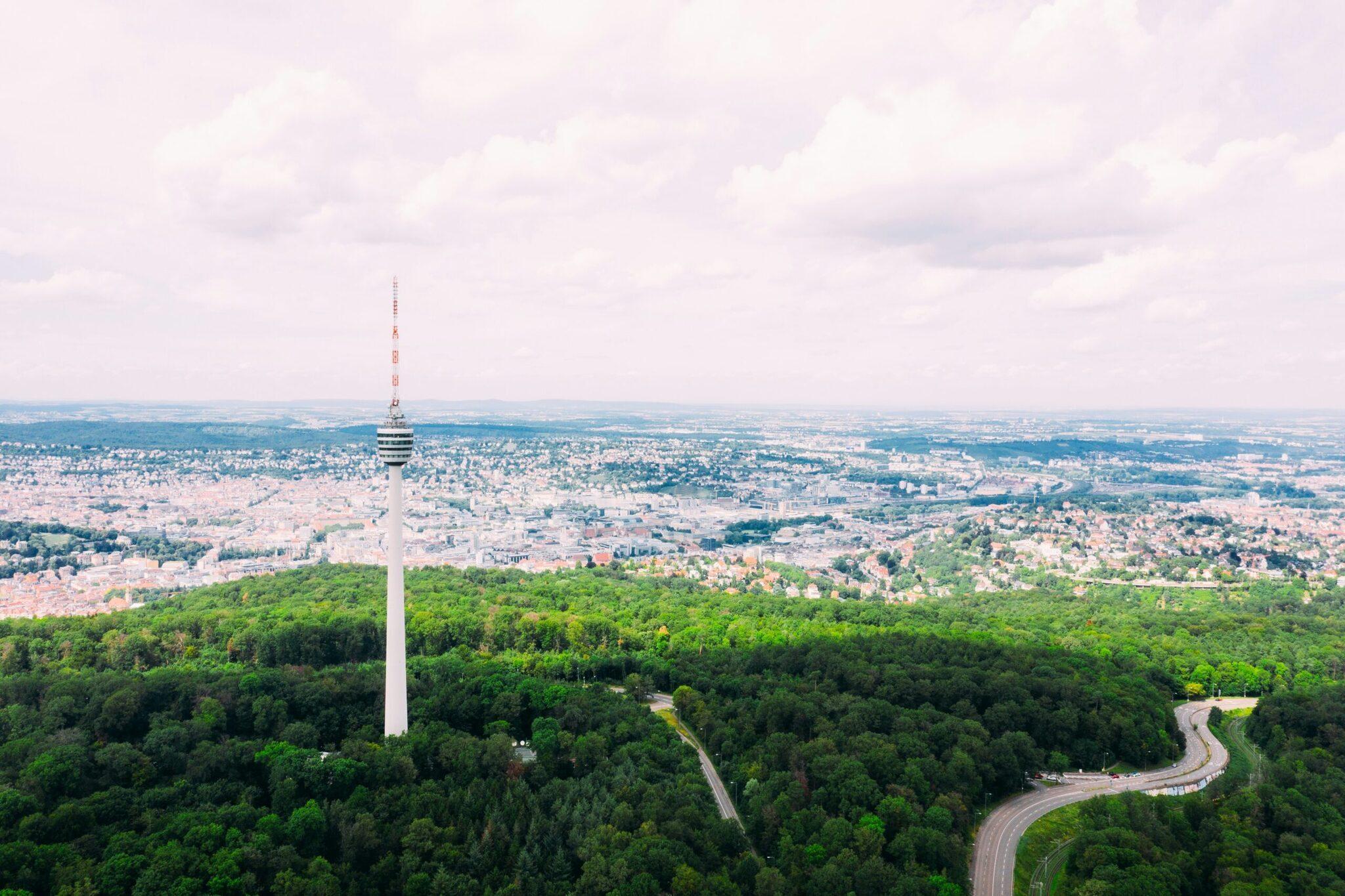 Aerial view of a white television tower dominating a dense forest with an expansive city skyline in the background
