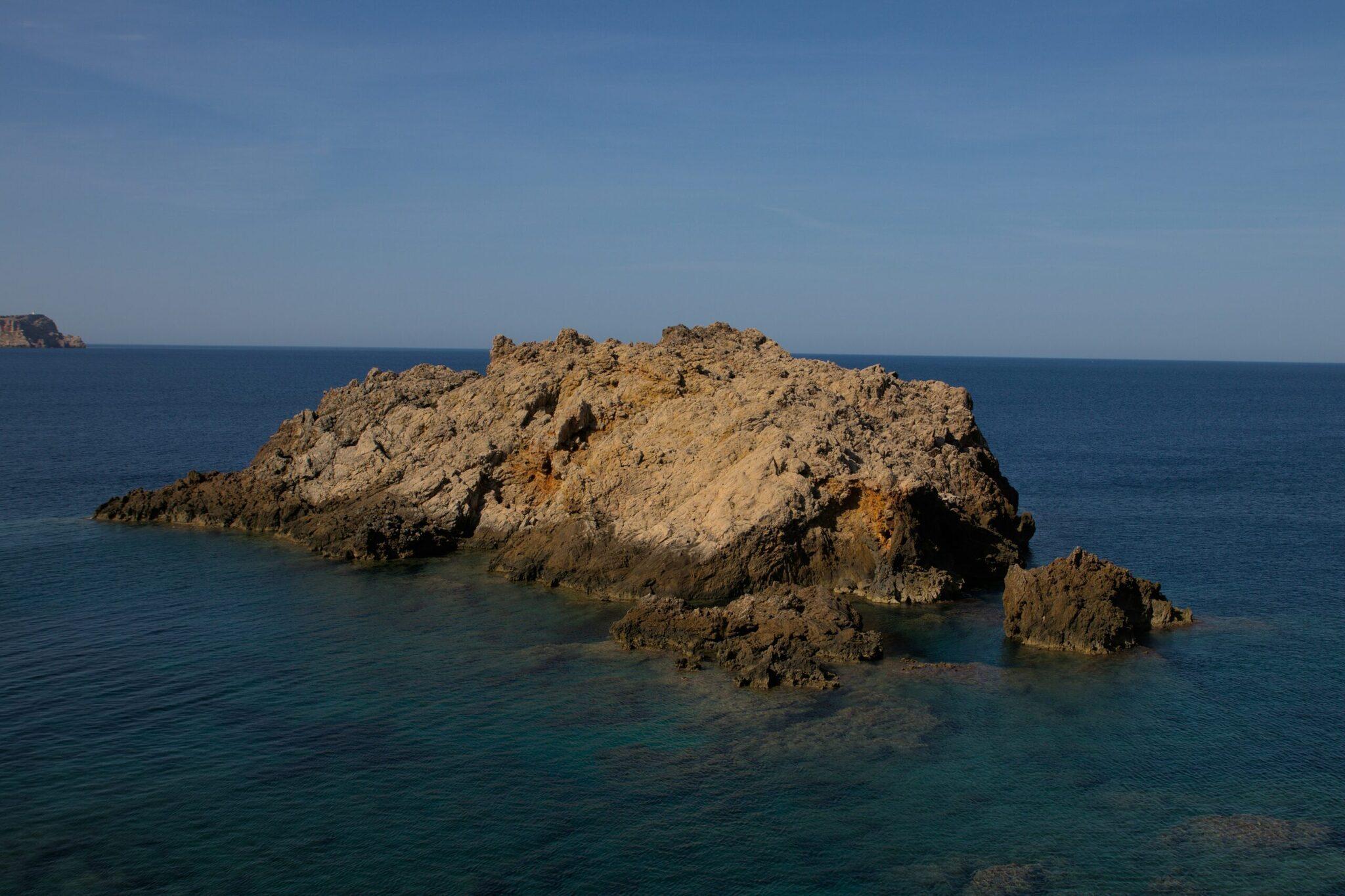 Islote rocoso con formaciones geológicas espectaculares que emerge del mar turquesa mediterráneo bajo un cielo azul despejado