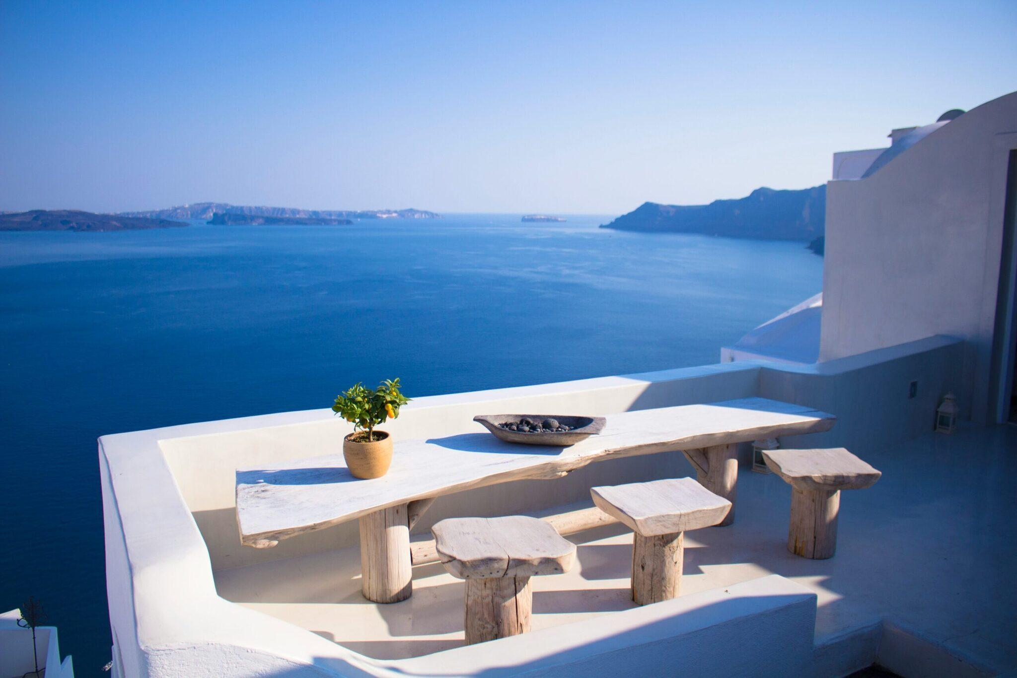 Terrasse blanche surplombant une baie méditerranéenne avec table en bois, stools et vue sur îles rocheuses