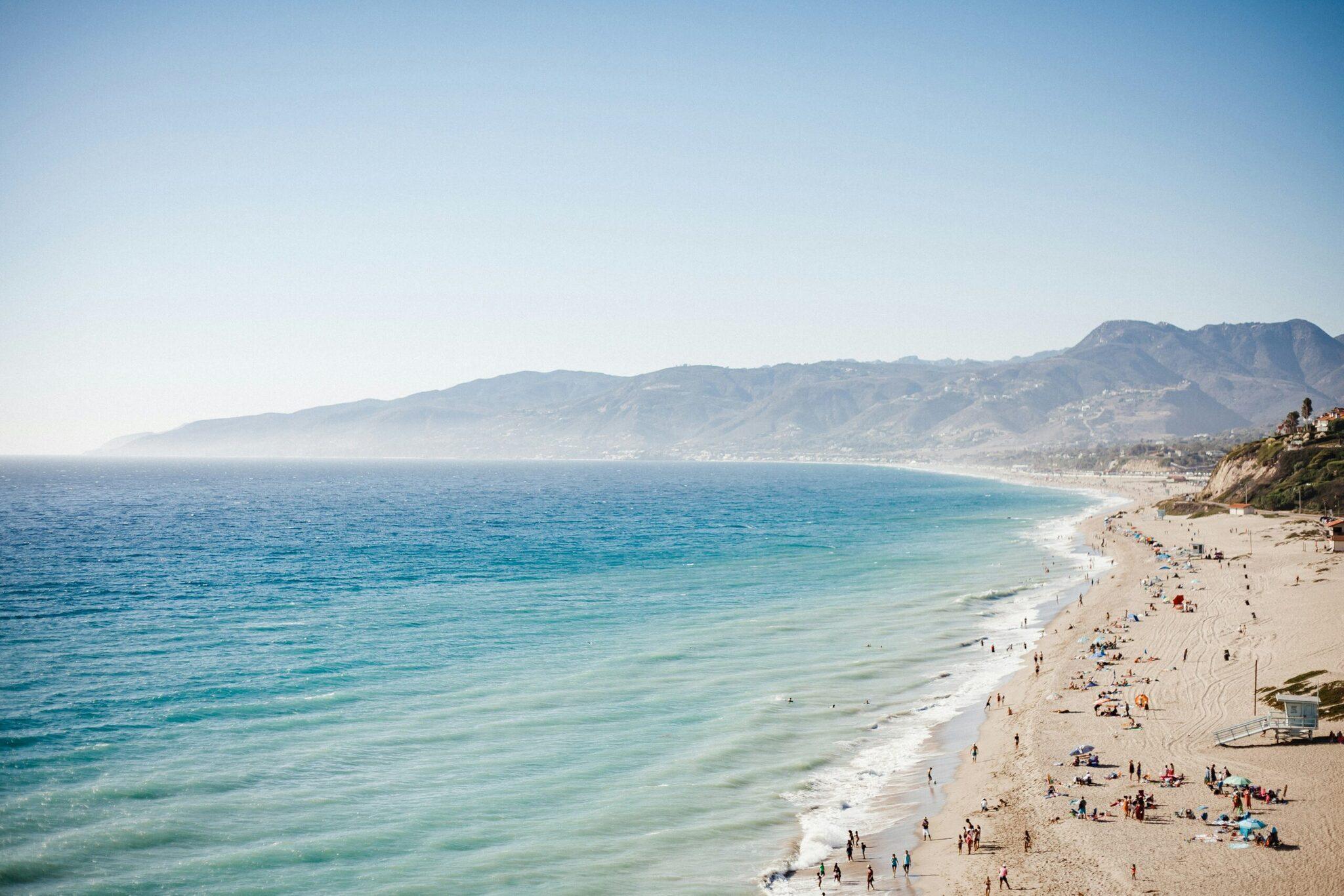 Ein langer Sandstrand mit Badegästen vor einem türkisfarbenen Ozean und bewaldeten Küstenbergen im Hintergrund