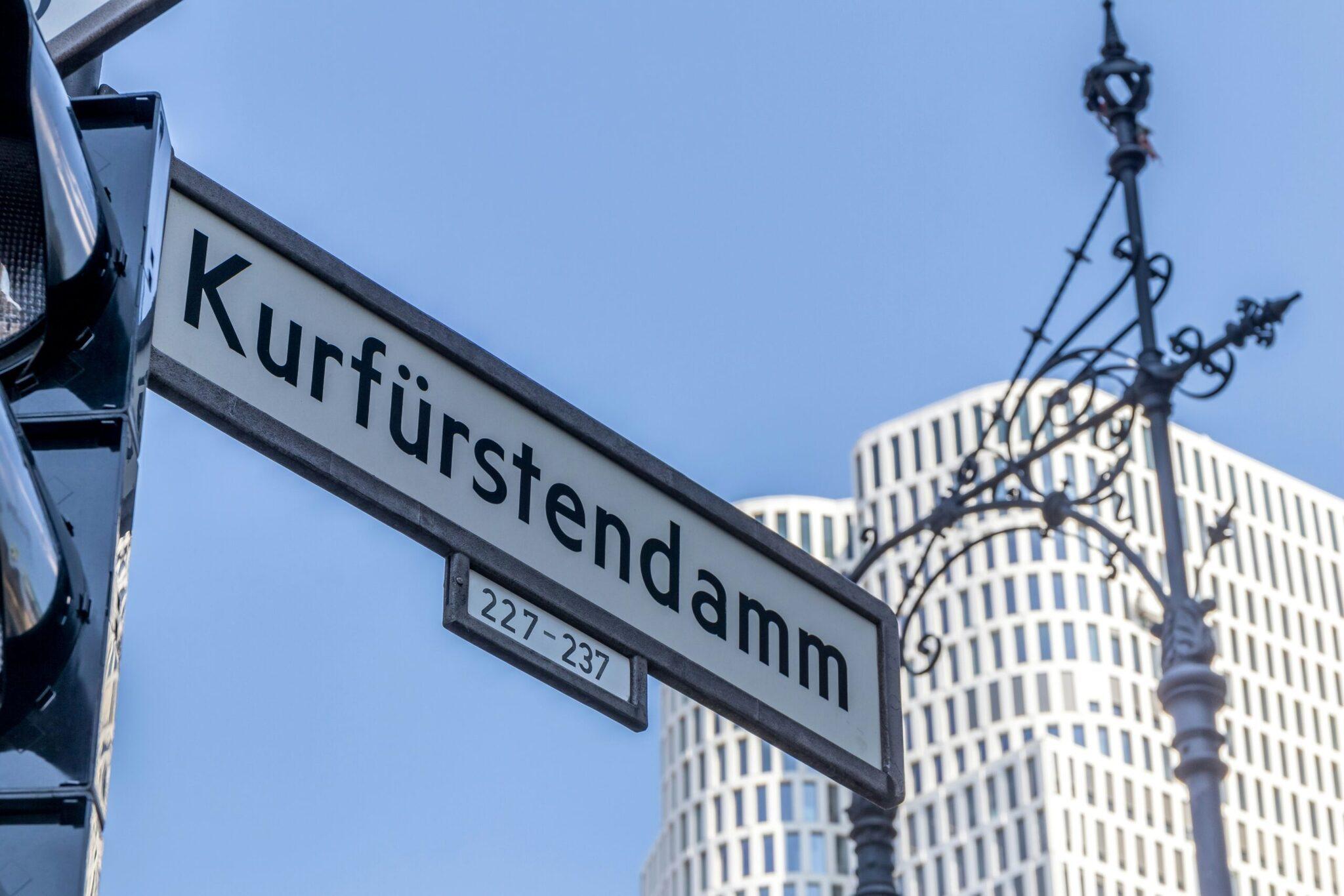 A Kurfürstendamm street sign with numbers 227-237 and modern Berlin buildings in the background under a blue sky