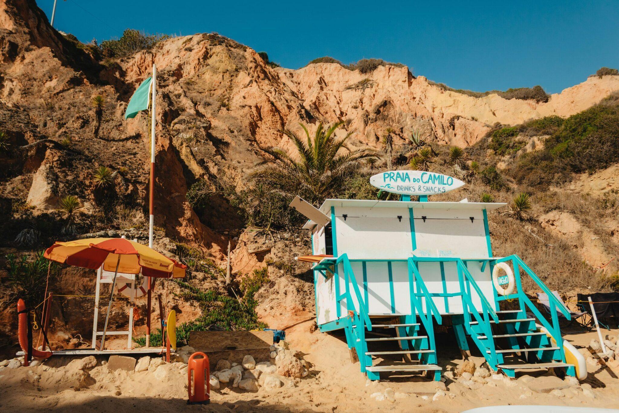 Ein farbenfroher kleiner Strandkiosk mit sonniger Terrasse, eingebettet am Fuß ockerfarbener Klippen in einer einsamen Küstenbucht