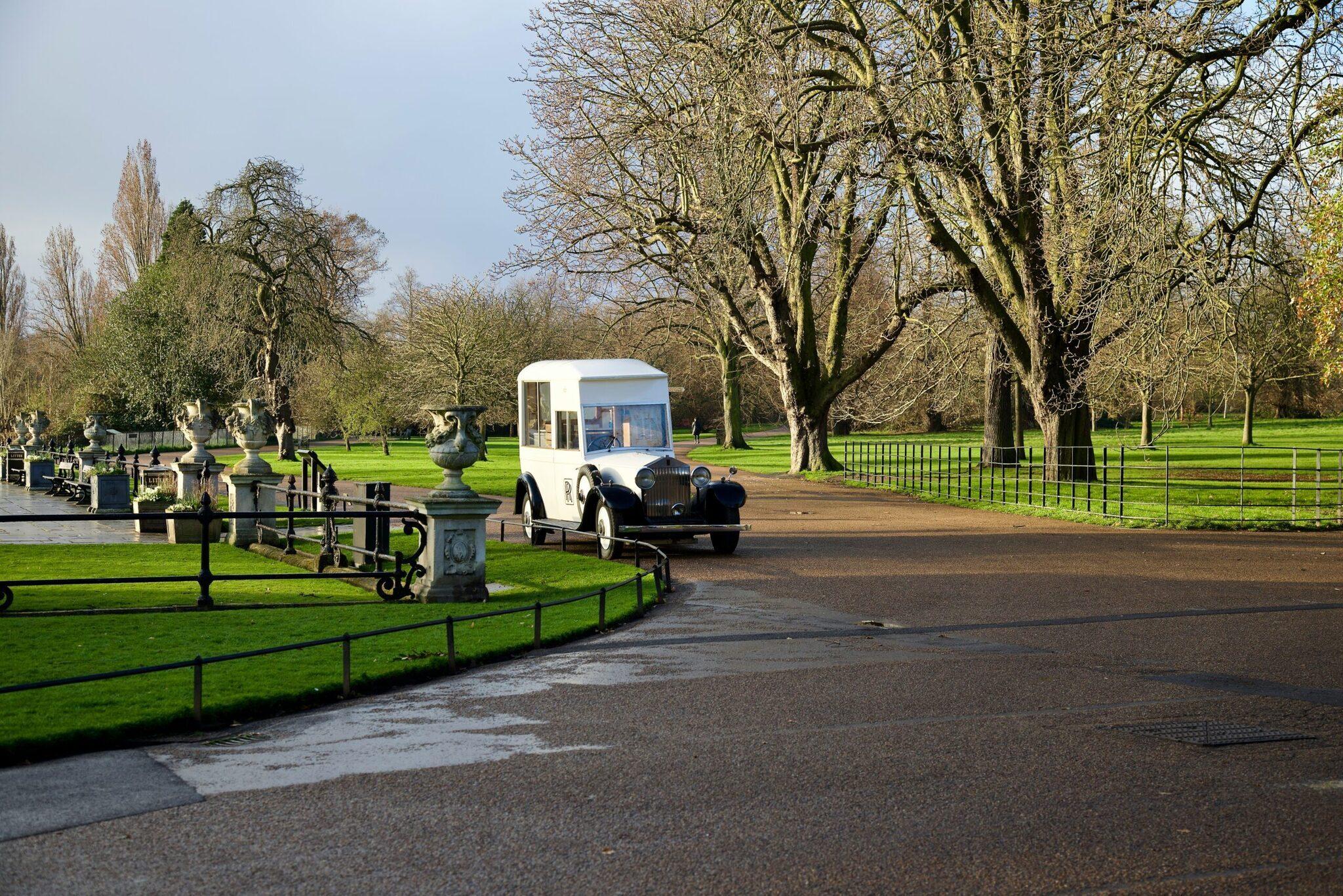 Ein schwarz-weißes Hochzeitsoldtimer-Auto fährt durch einen grünen Stadtpark mit jahrhundertealten Bäumen