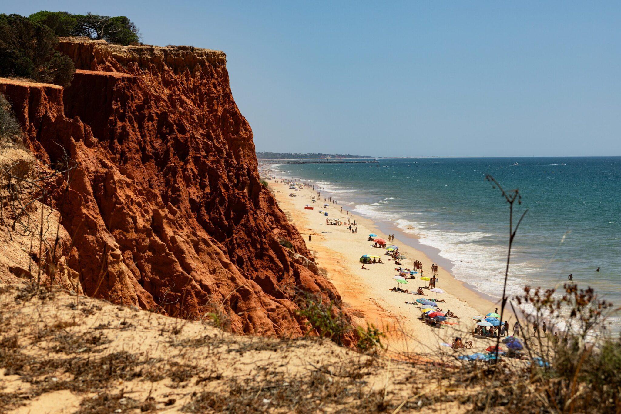 Ockerfarbene Klippe über einem goldenen Sandstrand mit Badegästen und Sonnenschirmen unter einem wolkenlosen blauen Himmel