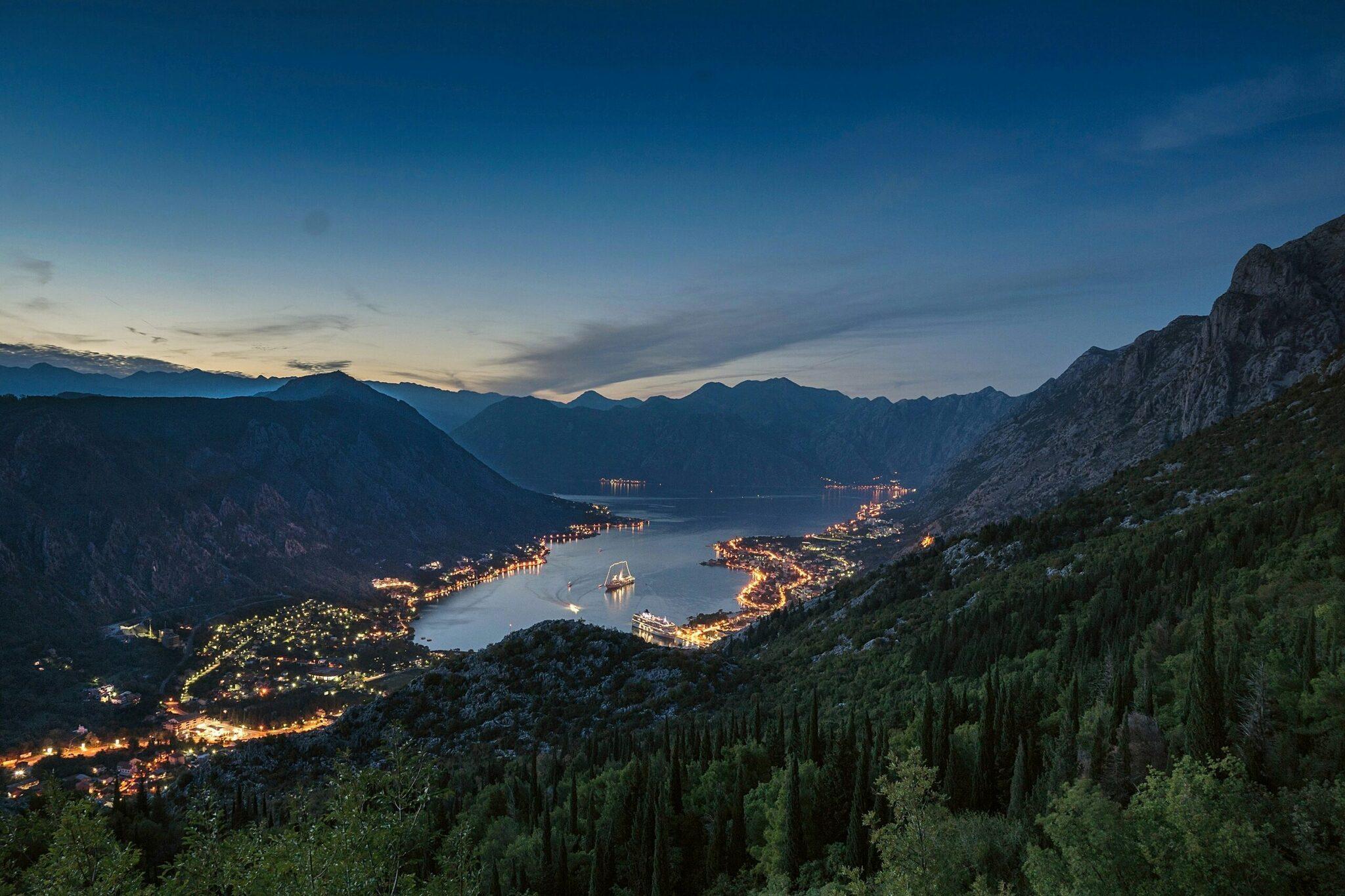 Vue nocturne d’une baie côtière encadrée de montagnes avec villes illuminées et voilier au crépuscule