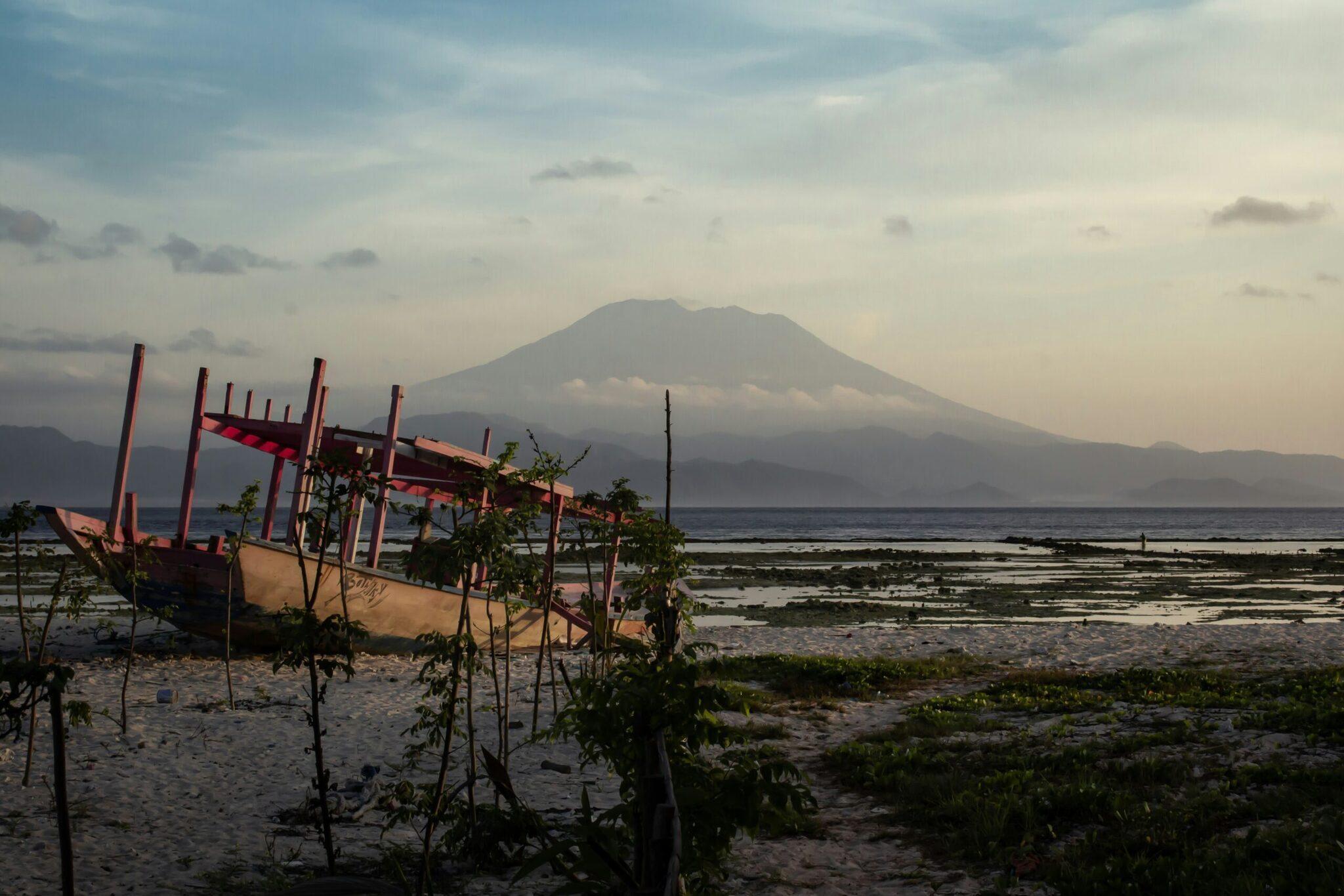Bateau de pêche rouge échoué sur une plage de sable avec un volcan majestueux en arrière-plan sous un ciel nuageux