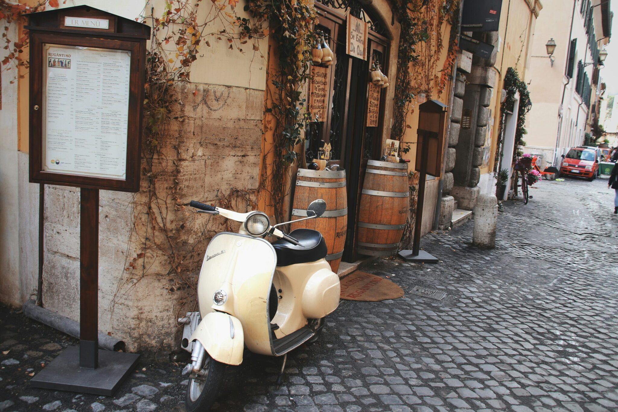 Un scooter Vespa blanc garé dans une ruelle pavée d’une vieille ville européenne avec façades ocre et lierre
