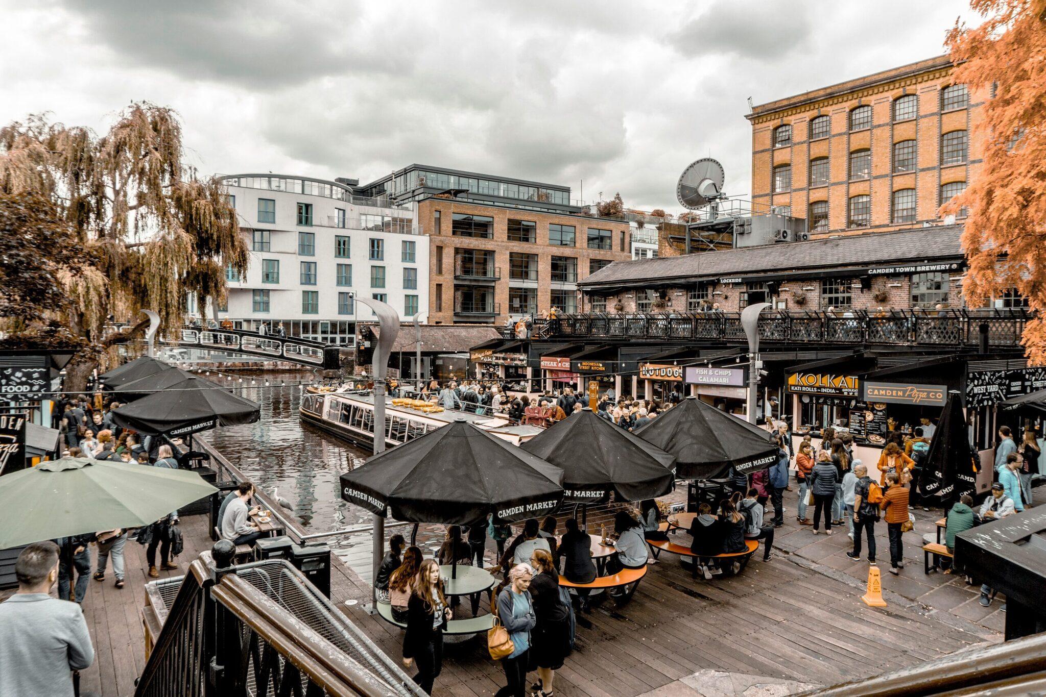 Ein belebter Markt am Kanalwasser mit Terrassen, Läden und modernen Gebäuden im Hintergrund bei bewölktem Wetter