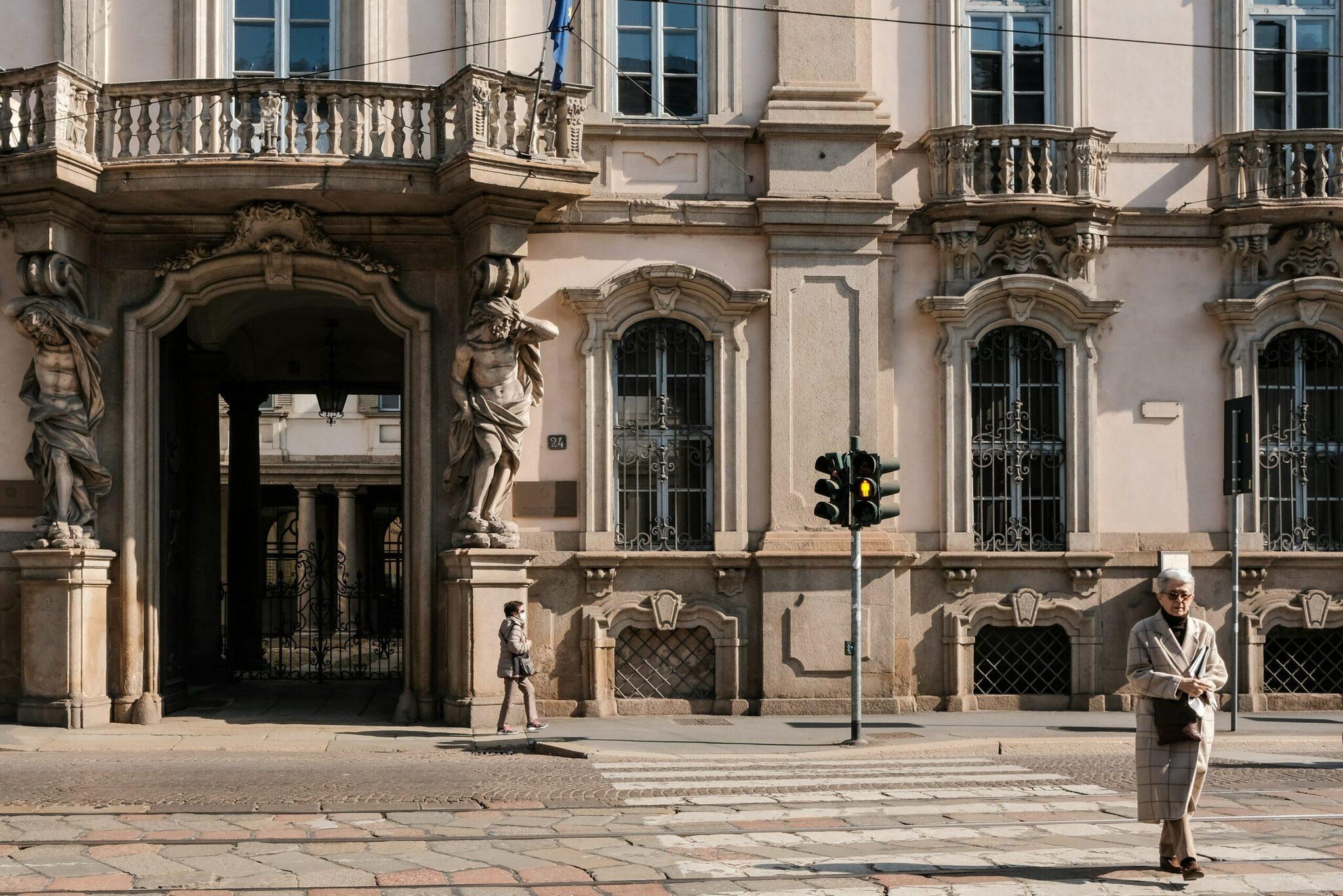Une place pavée devant un élégant palais Renaissance avec colonnes et sculptures, quelques piétons sous le soleil