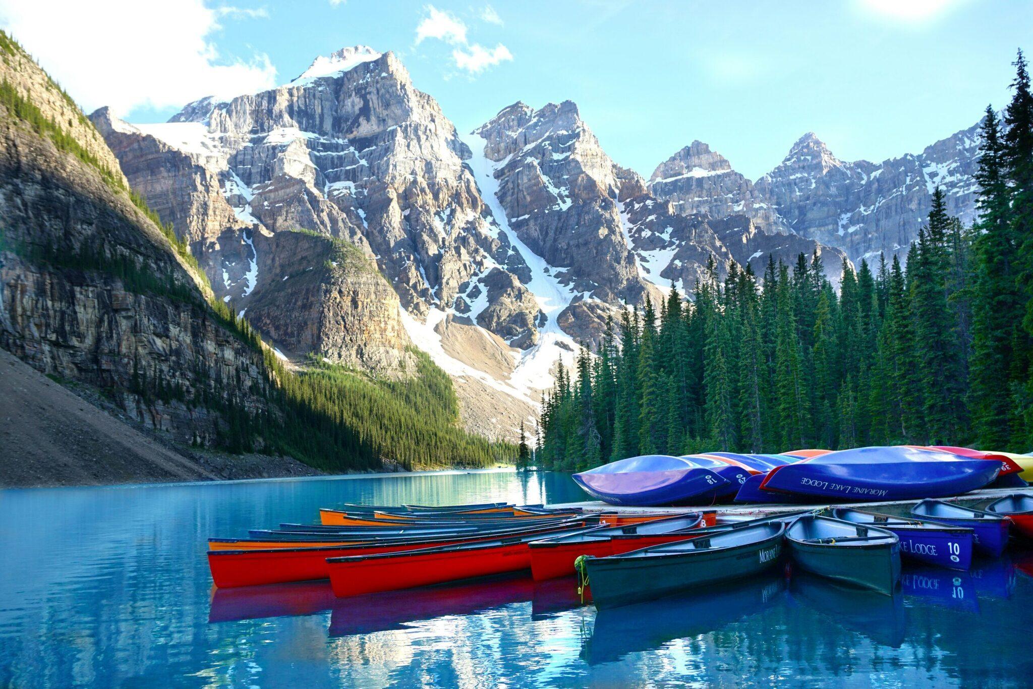 Des canoës colorés alignés sur un lac turquoise encadré de montagnes rocheuses enneigées et de forêts de conifères