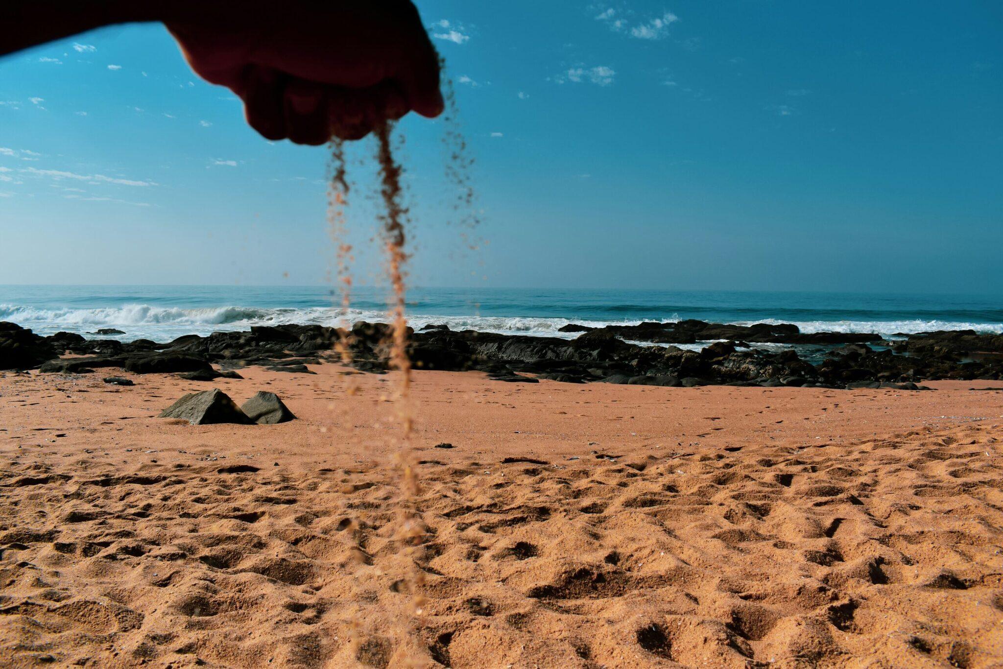 Une main verse du sable fin sur une plage rocheuse avec vue sur l’océan et les vagues