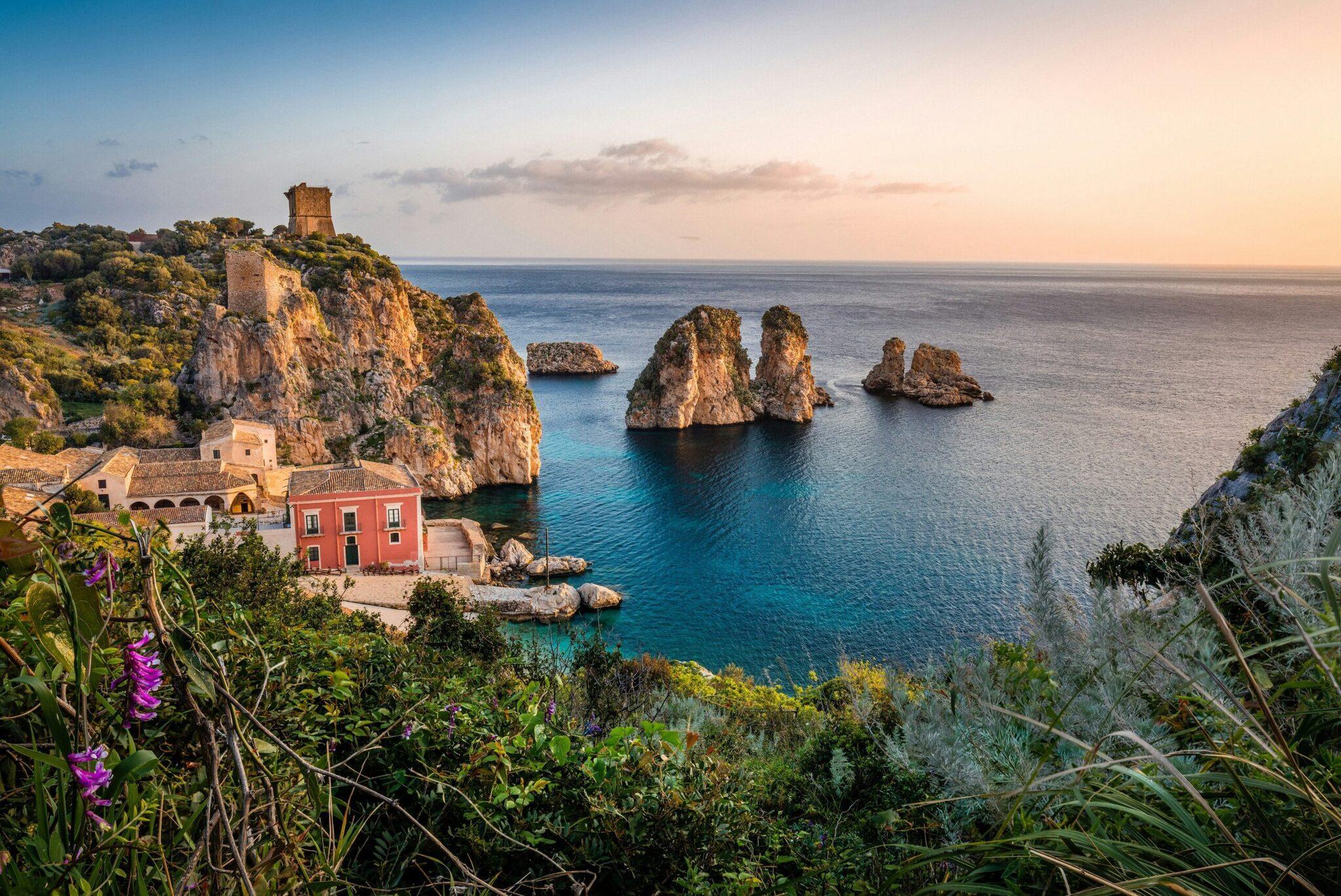 Vue côtière méditerranéenne avec maisons colorées, falaises rocheuses et formations géologiques en mer au coucher du soleil