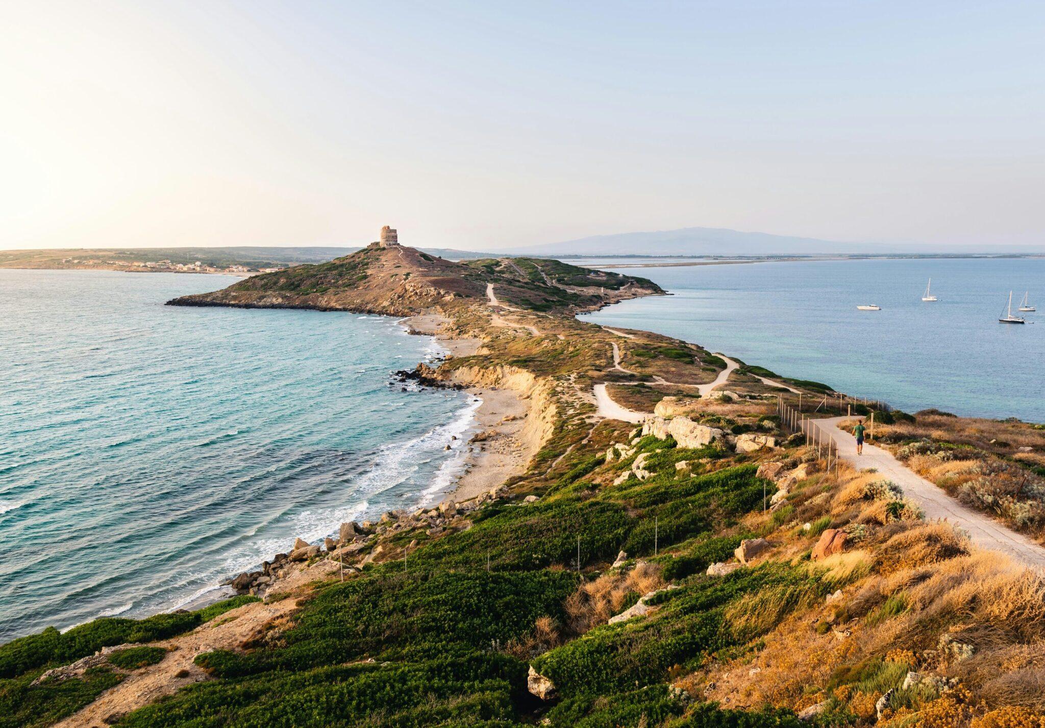 Sentier côtier sinueux longeant une baie turquoise avec îlot rocheux fortifié et voiliers ancrés