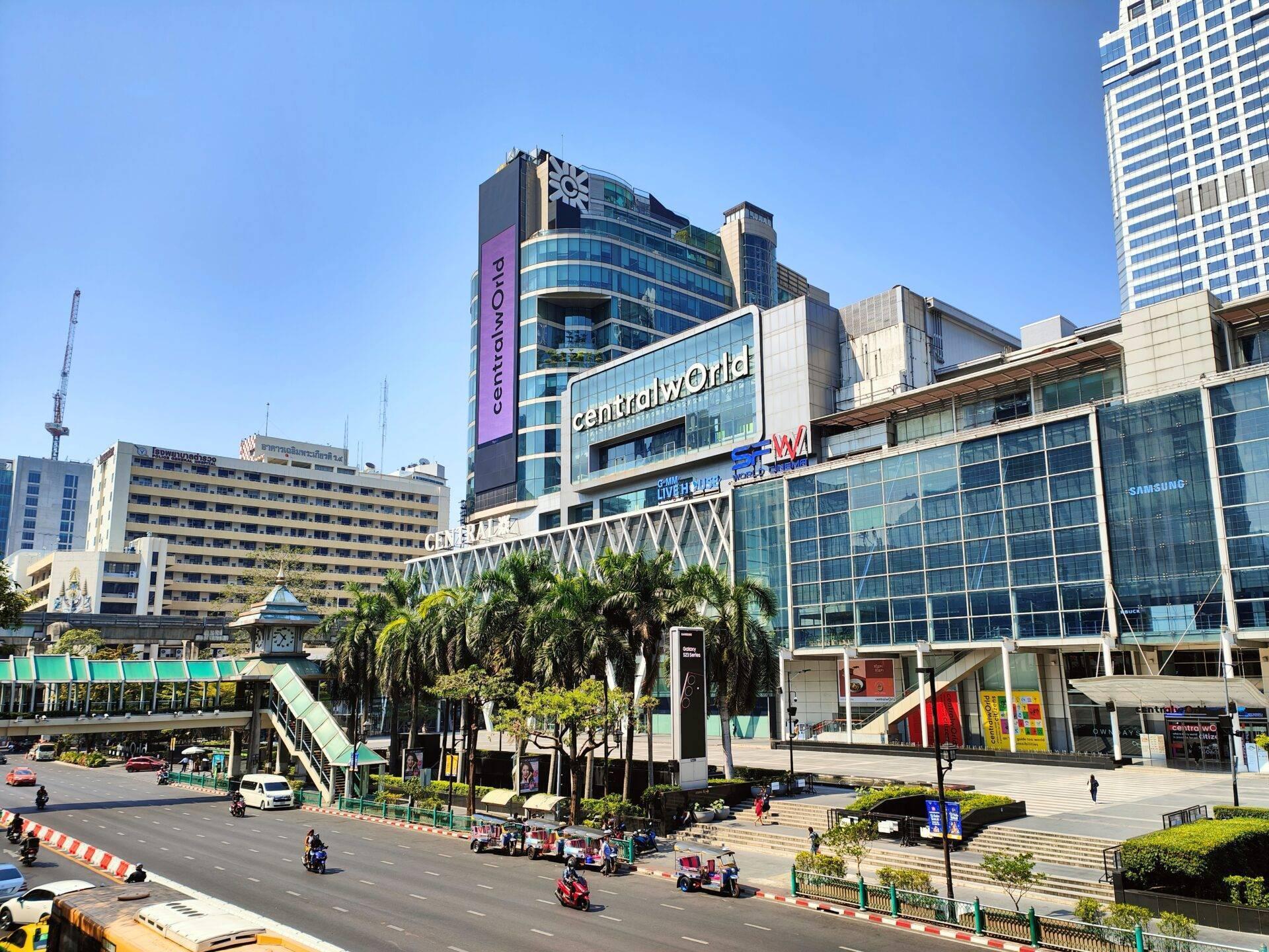 Une grande place commerciale urbaine avec centre commercial moderne, gratte-ciels, palmiers et tuk-tuks rouges sous un ciel bleu