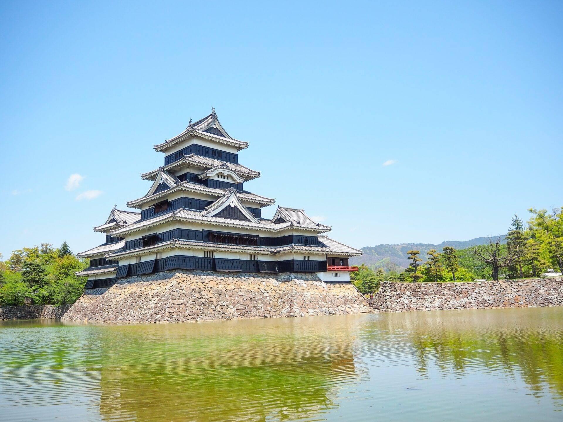 Un château japonais traditionnel avec toitures étagées surplombant un fossé rempli d’eau, entouré de verdure