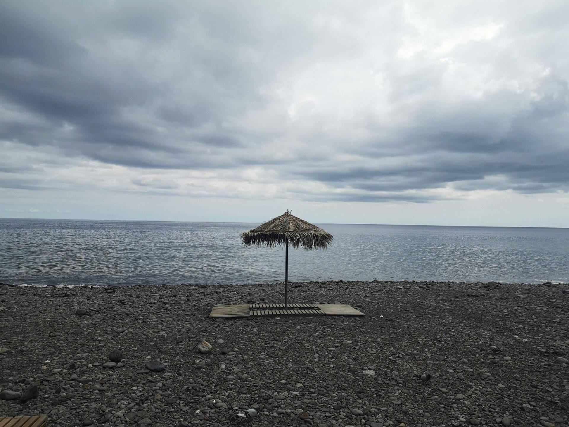 A solitary straw parasol on a grey pebble beach facing a calm sea under a cloudy sky