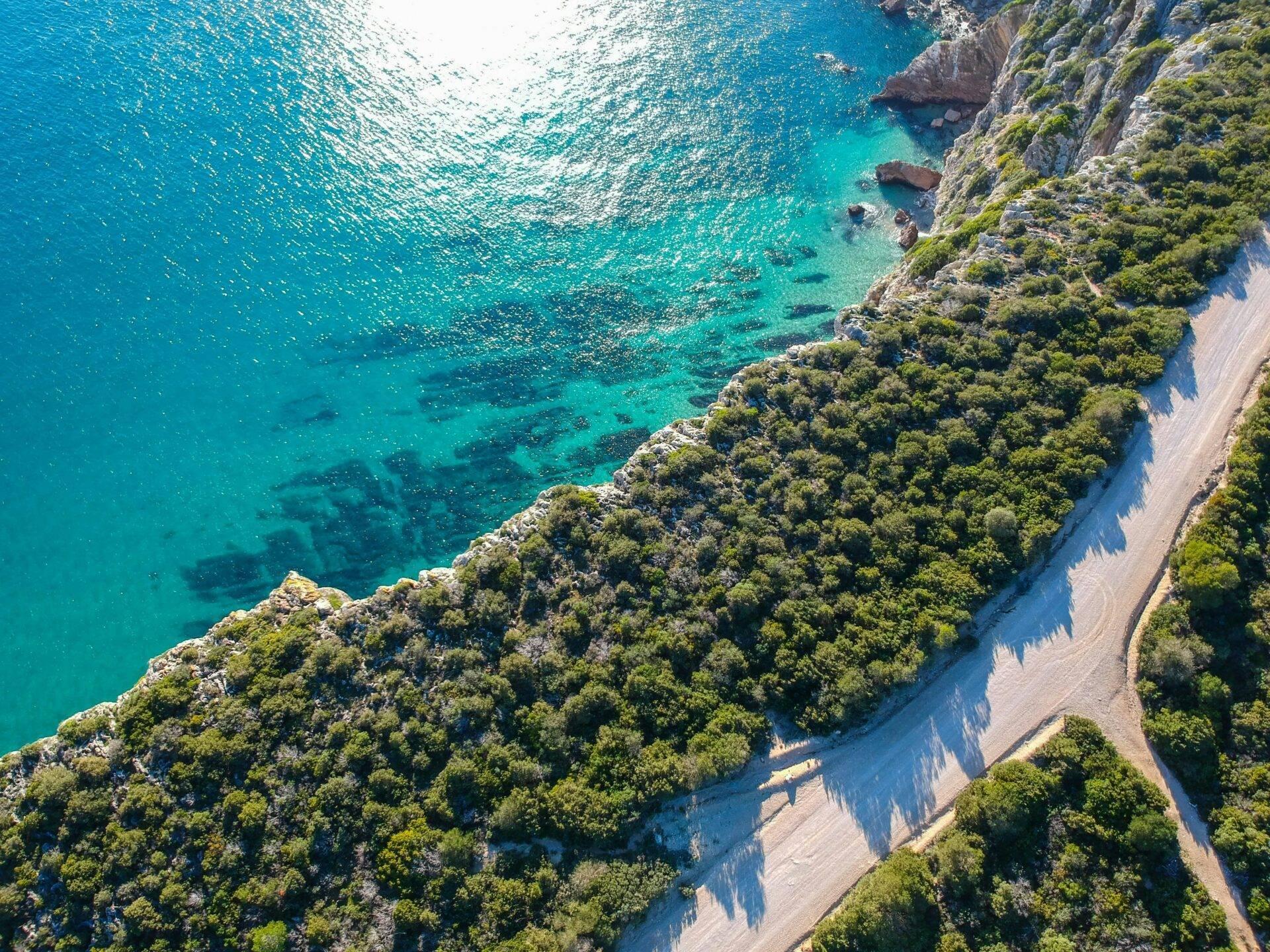 Vista aerea di una costa mediterranea con scogliere boscose, spiaggia di sabbia e acque turchesi cristalline