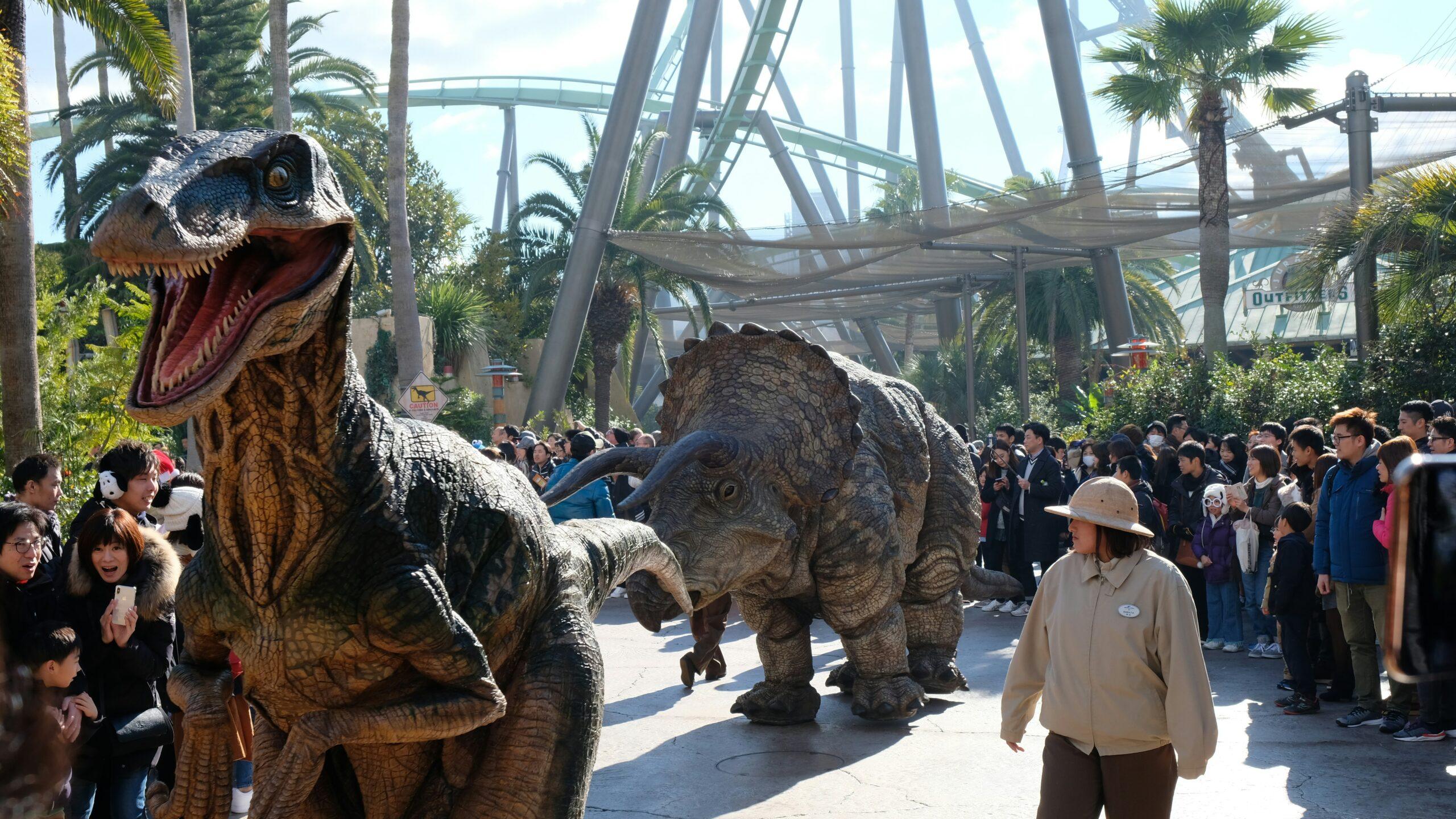 Défilé de dinosaures animatroniques dans un parc à thème avec foule de visiteurs et structures métalliques de montagnes russes en arrière-plan