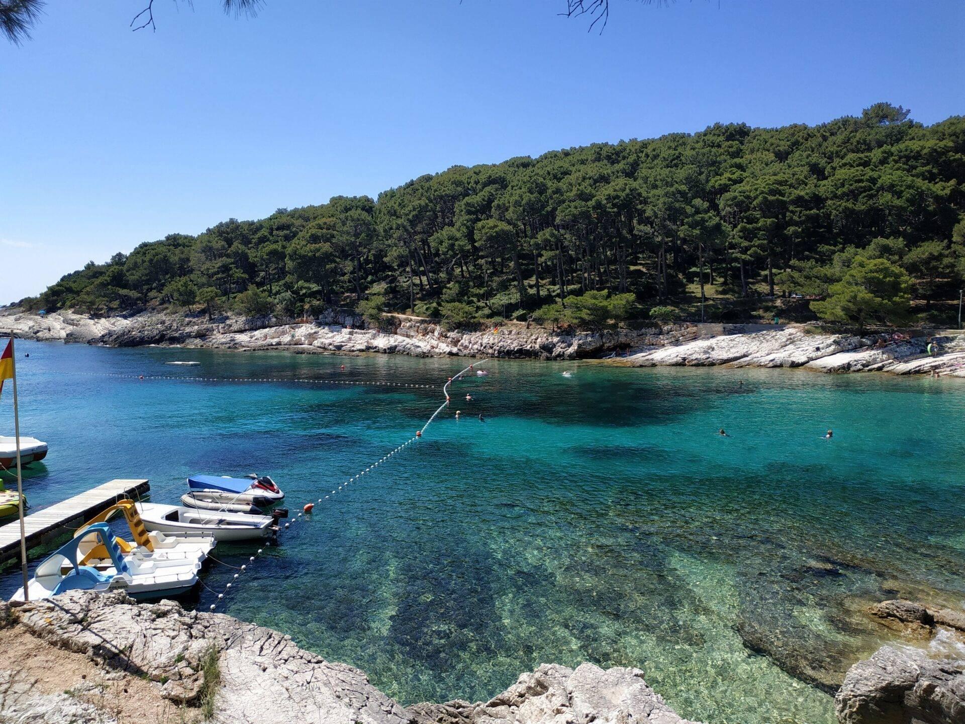 Mediterrane Bucht mit türkisfarbenem Wasser, vertäuten Booten, Felsklippen und Pinienwald