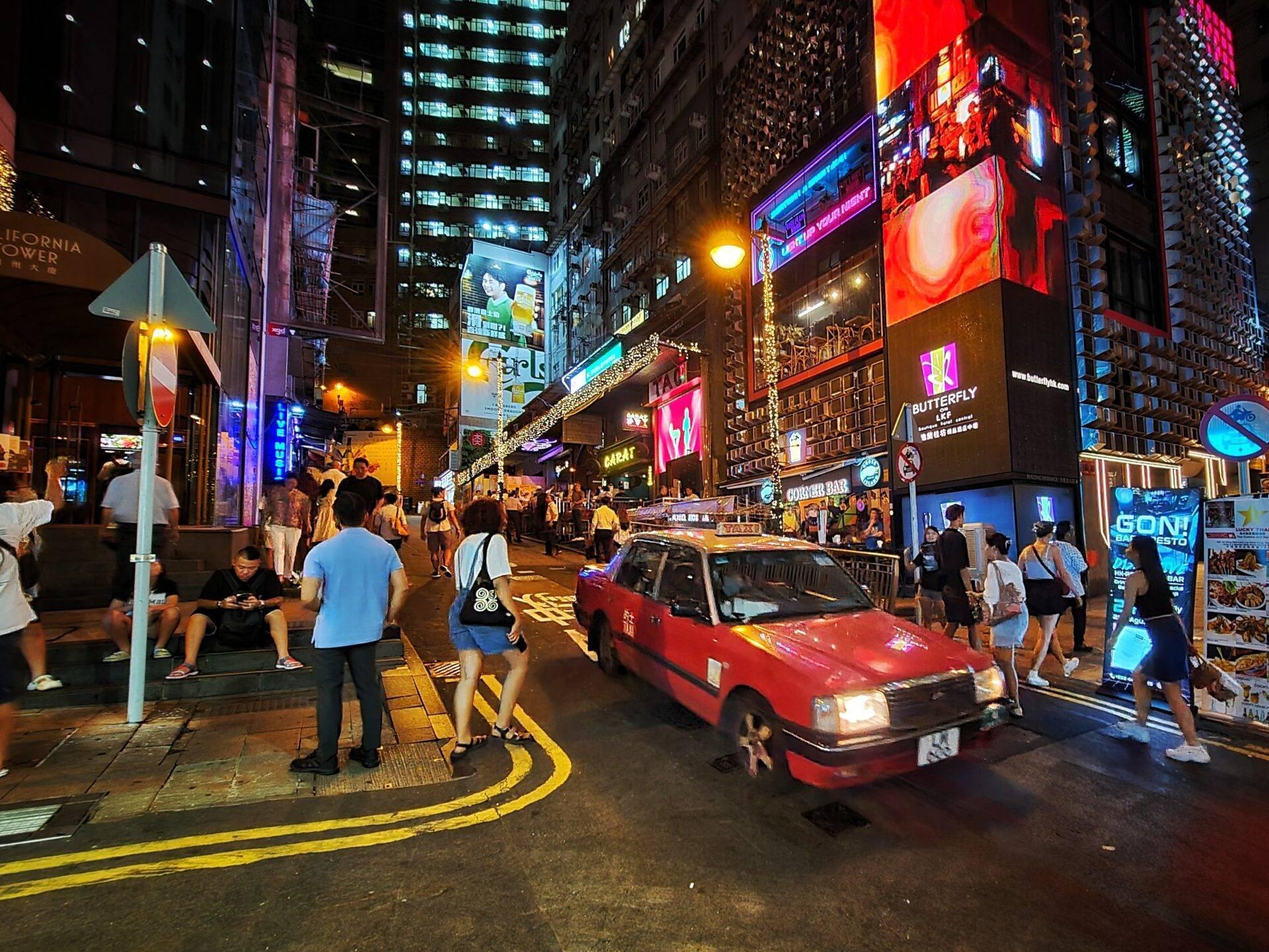 Une rue piétonne animée la nuit avec gratte-ciel illuminés, panneaux publicitaires colorés et taxi rouge circulant parmi les passants