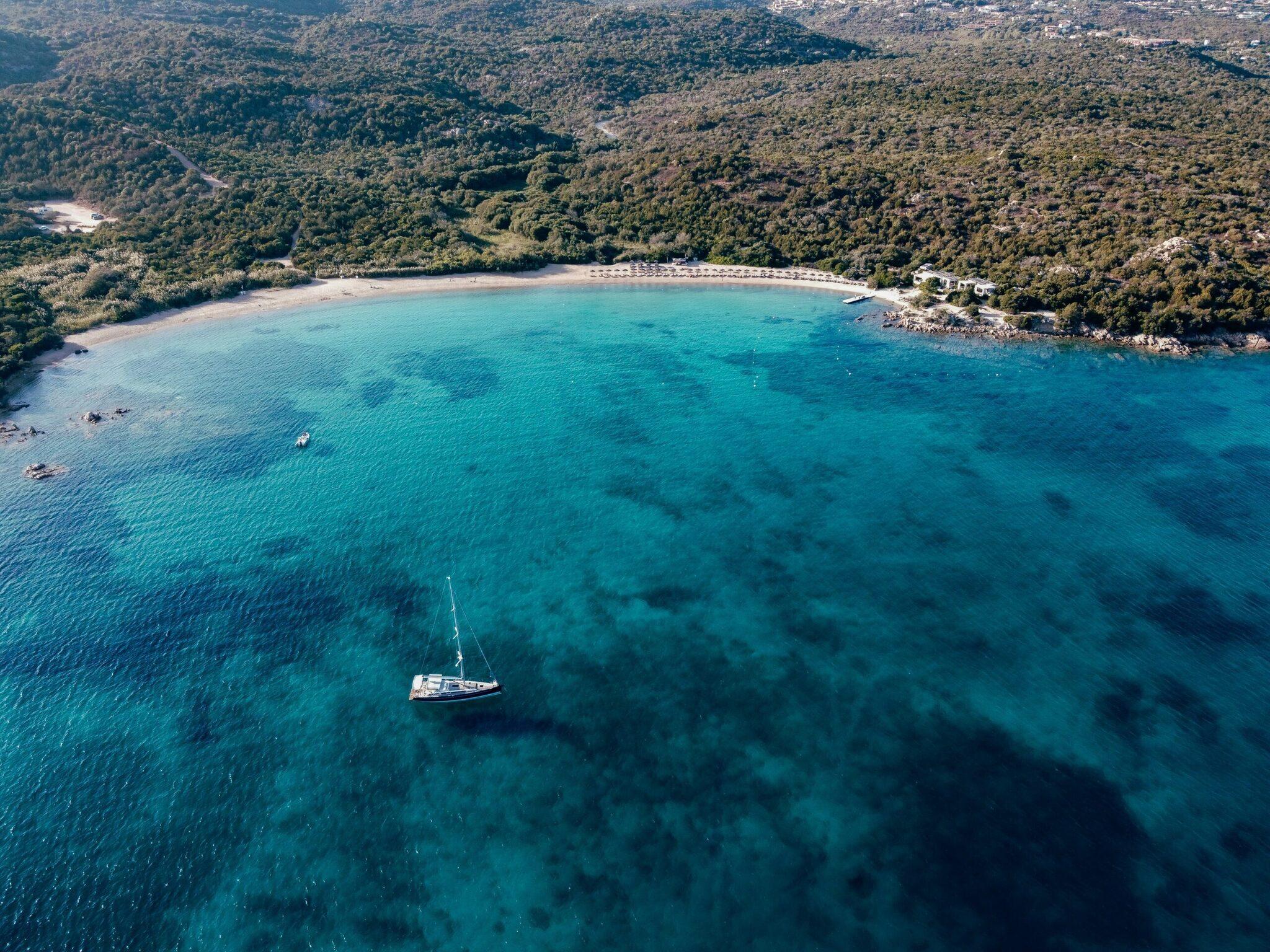 Vue aérienne d’une baie méditerranéenne aux eaux turquoise avec voilier, plage de sable et forêts côtières