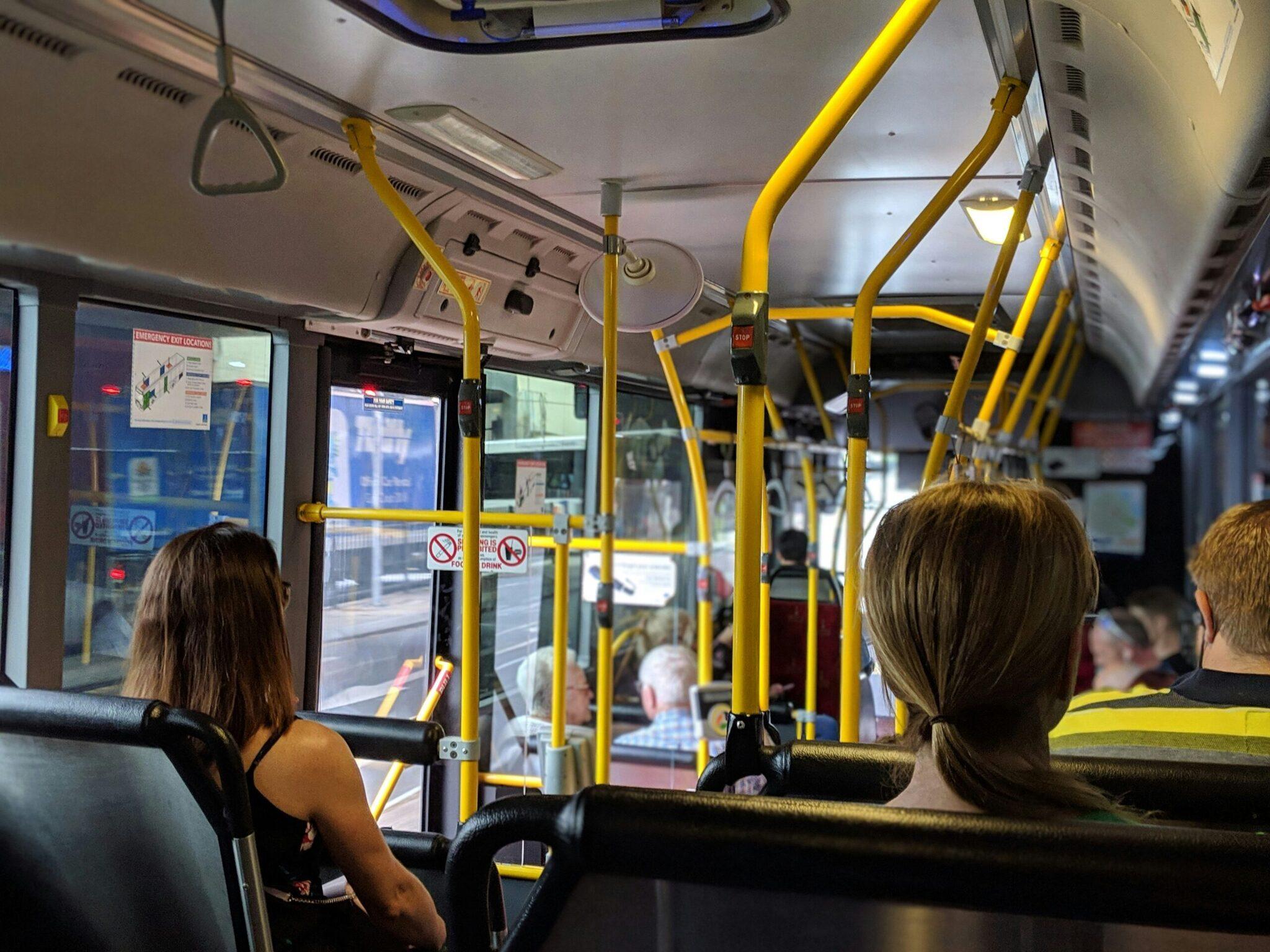 Intérieur d’un bus avec passagers assis, barres de maintien jaunes et vitres teintées donnant sur la rue