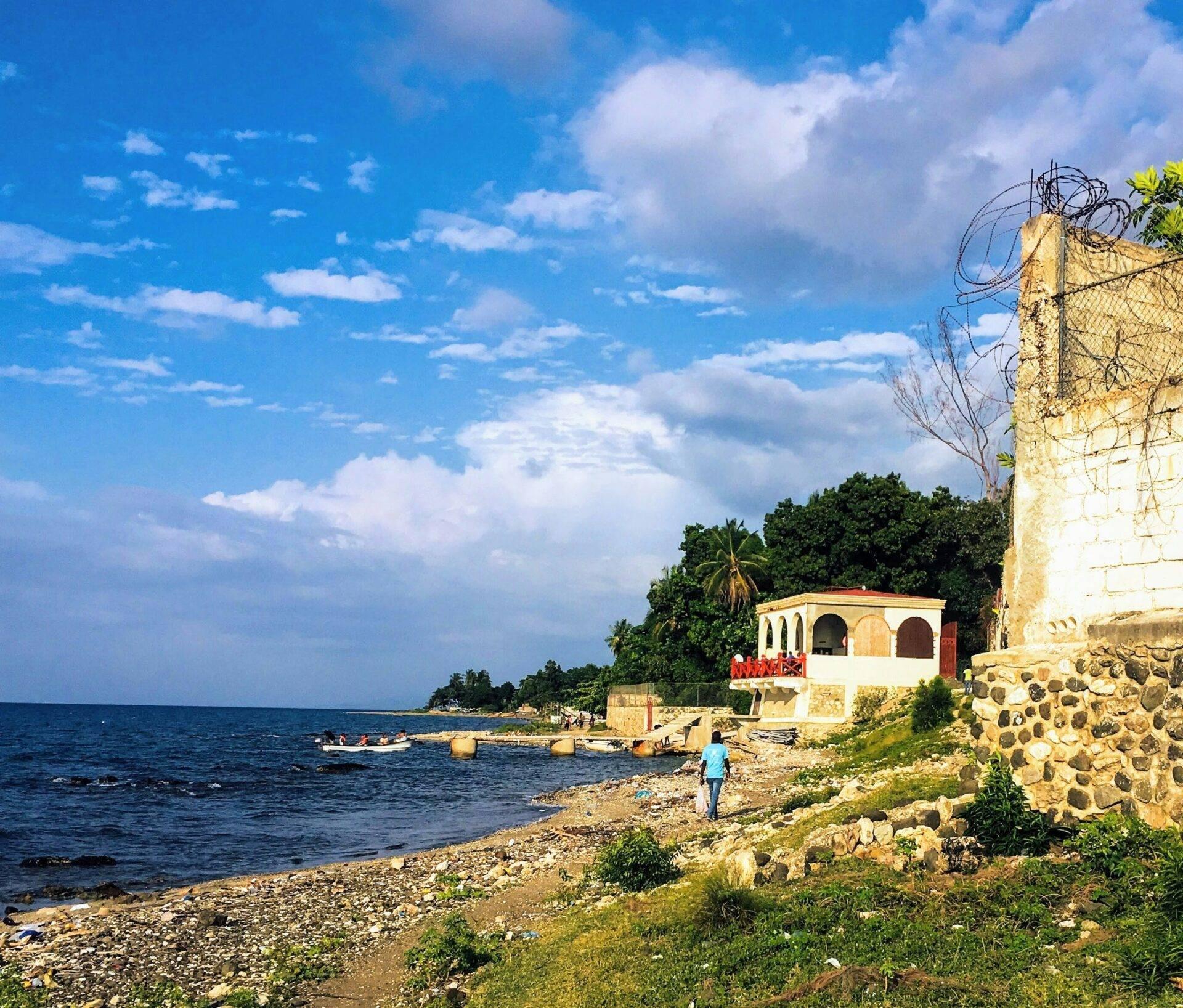 Una persona cammina su una spiaggia rocciosa di fronte a una casa colorata in pietra sul bordo del mare con barche