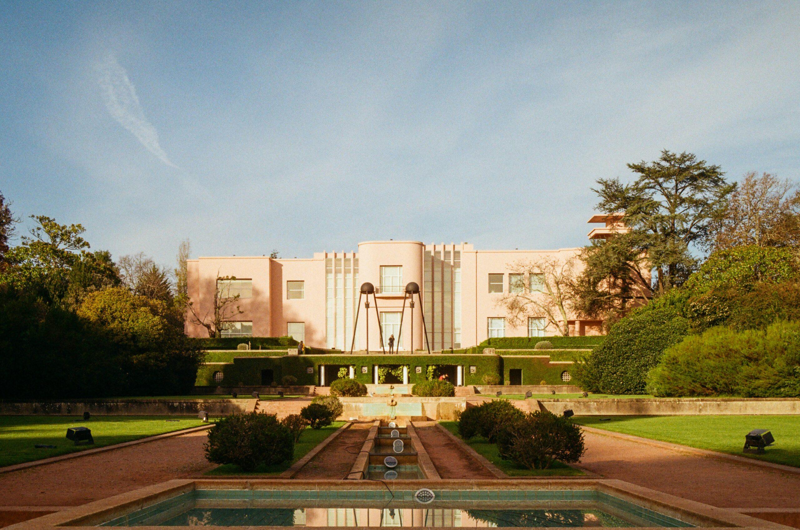 A pale pink Art Deco building with fountains and a reflecting pool, surrounded by lush gardens and trees