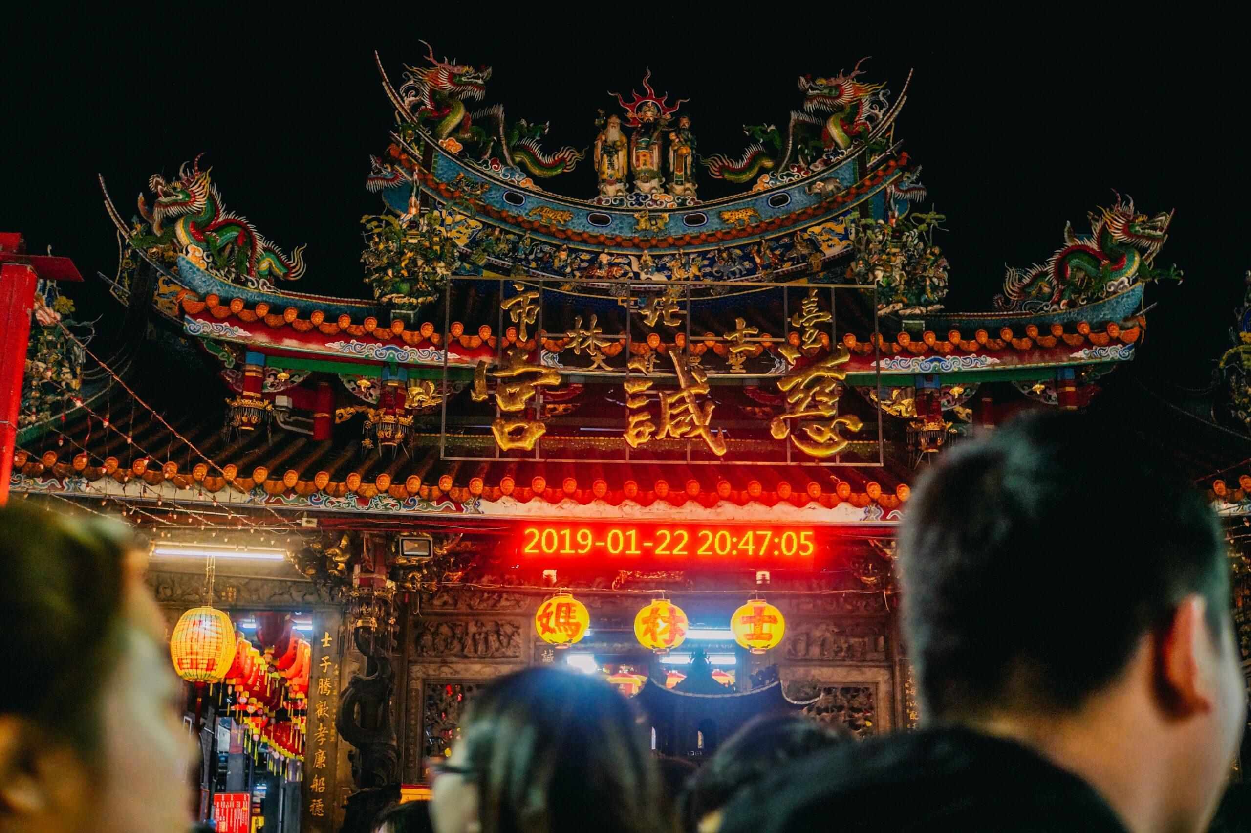 Un temple asiatique richement orné illuminé la nuit avec lanternes dorées et dragons sculptés, observé par des visiteurs