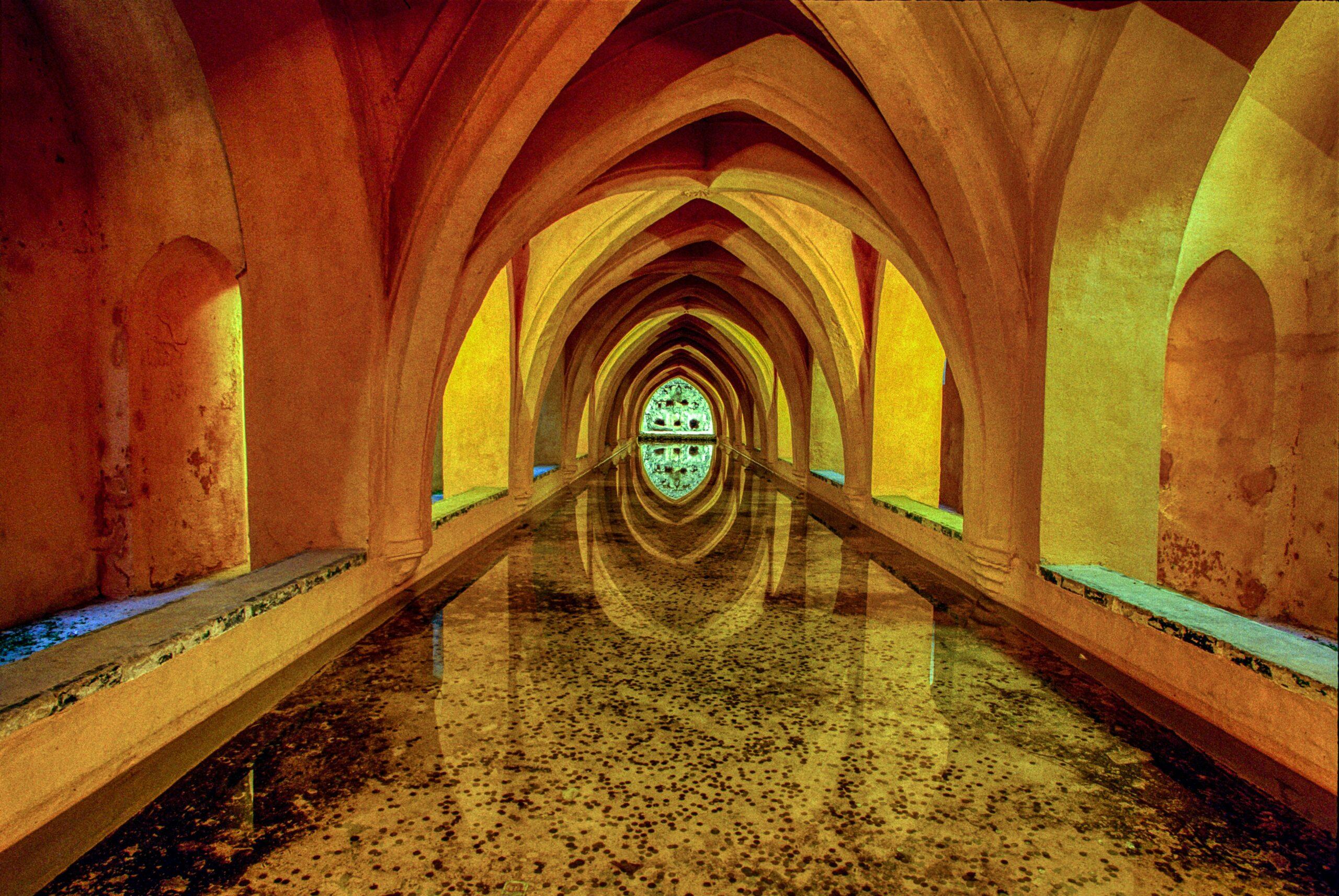 Galleria sotterranea con volte gotiche illuminate in giallo e rosso, pavimento allagato che riflette l’architettura