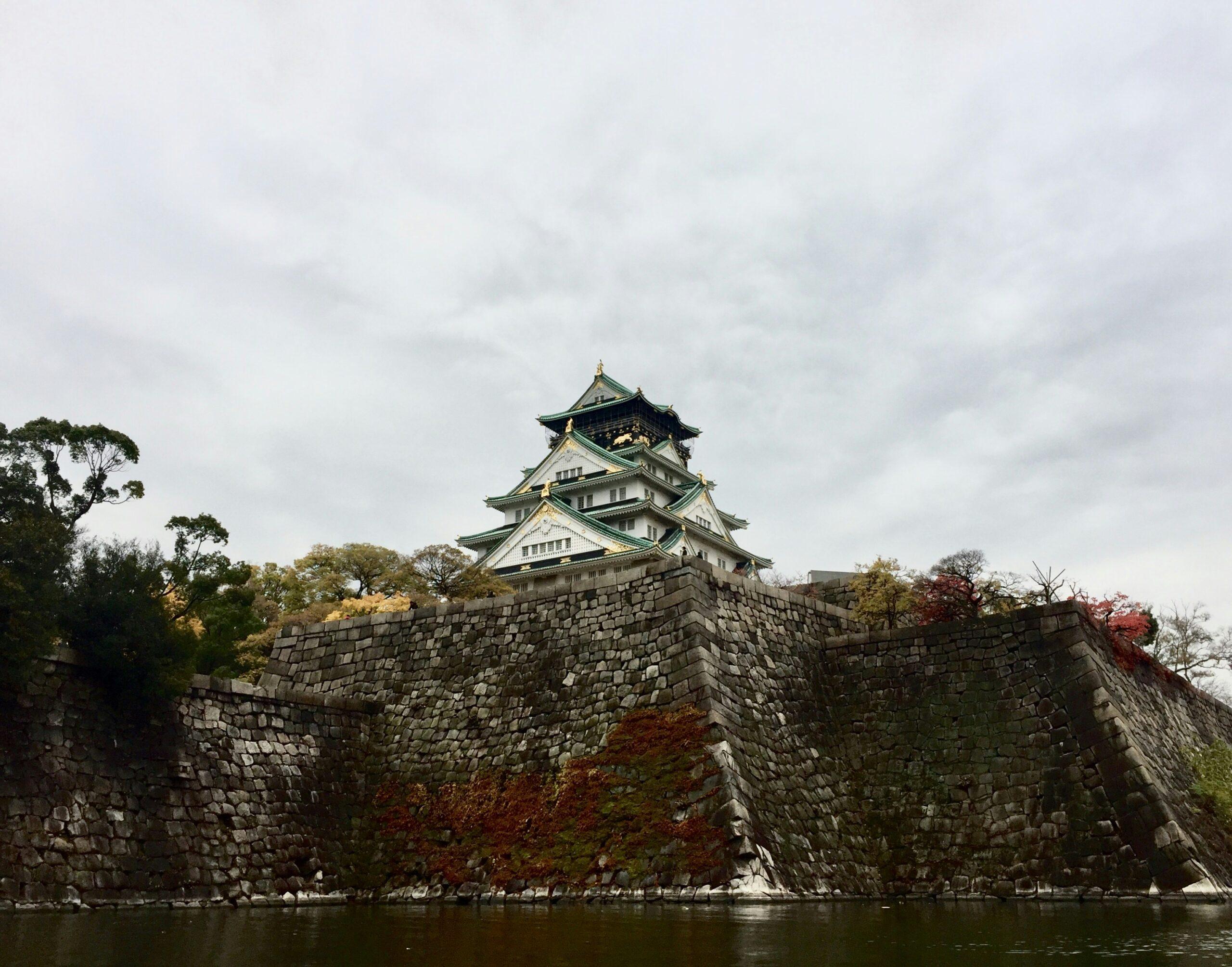 Un château japonais aux toits verts perchés sur d’imposants murs de pierre, surplombant l’eau avec végétation d’automne