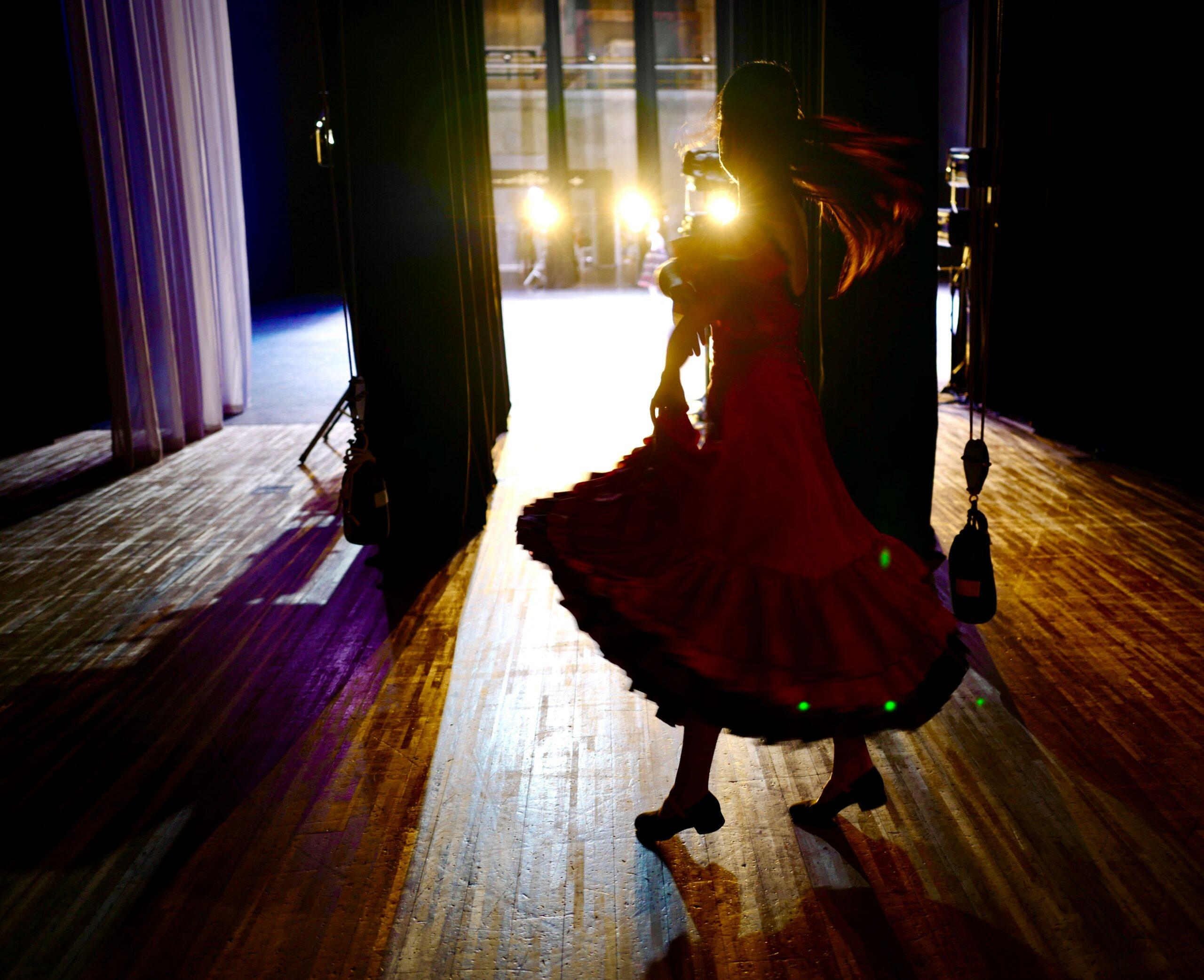 Una donna in abito rosso danza sul palco, illuminata dai riflettori in una sala da spettacolo
