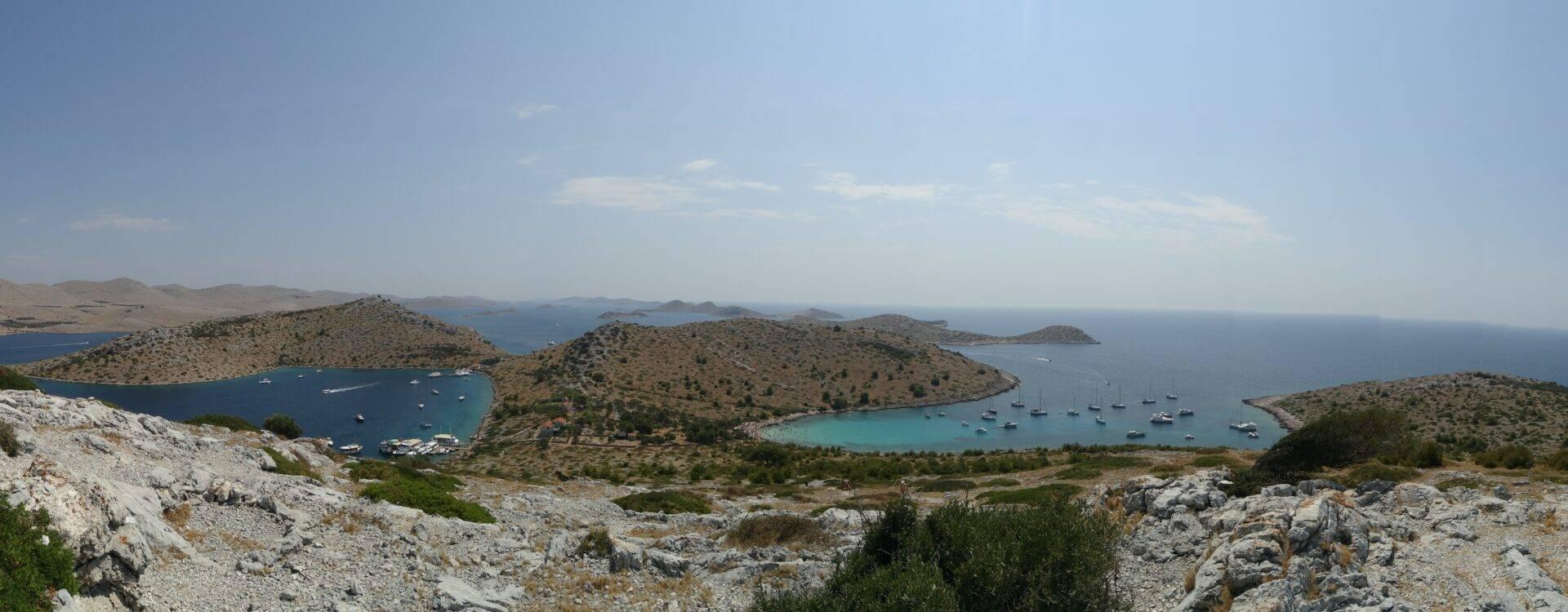 Panoramaansicht einer mediterranen Bucht mit bewaldeten Inseln, türkisfarbenem Wasser und verankerten Segelbooten
