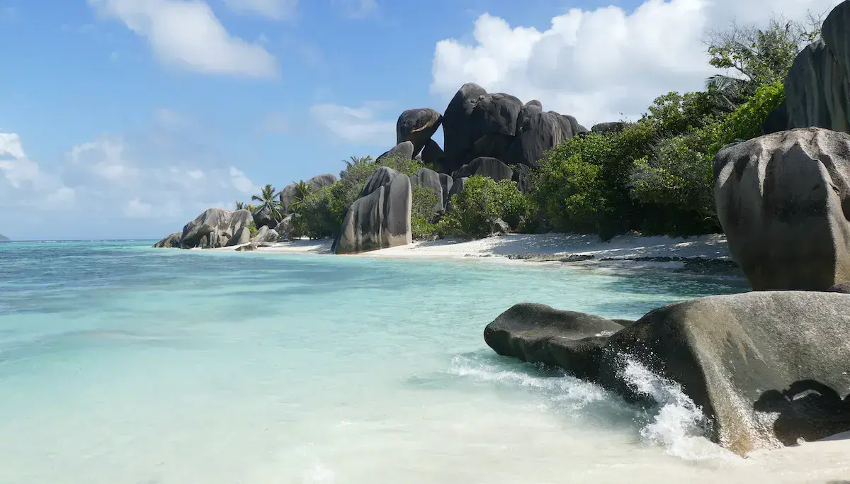 Plage tropicale avec rochers granitiques massifs, sable blanc et eau turquoise cristalline sous ciel dégagé