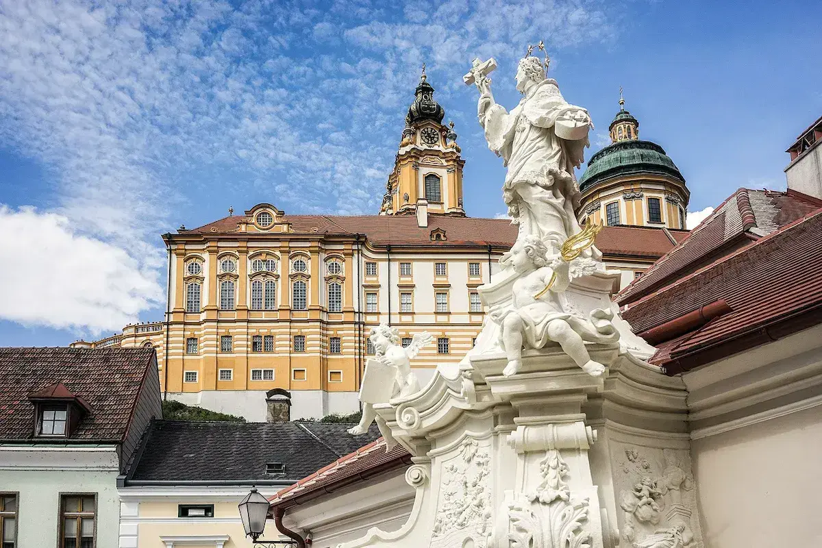 Statue baroque blanche devant un monastère jaune aux tours dorées sous ciel bleu nuageux