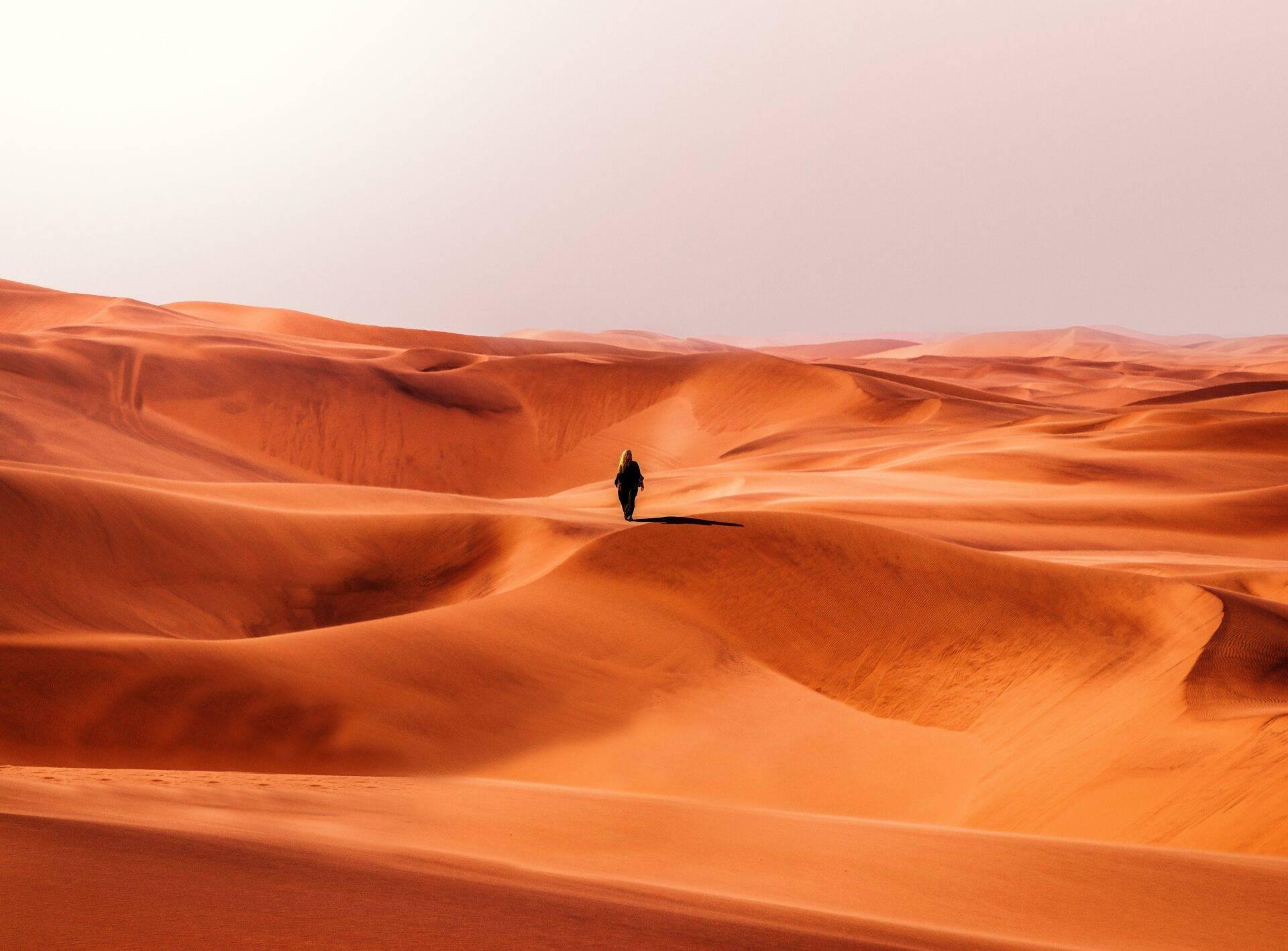 Un voyageur solitaire marche dans les dunes de sable orange d’un désert, sous un ciel dégagé
