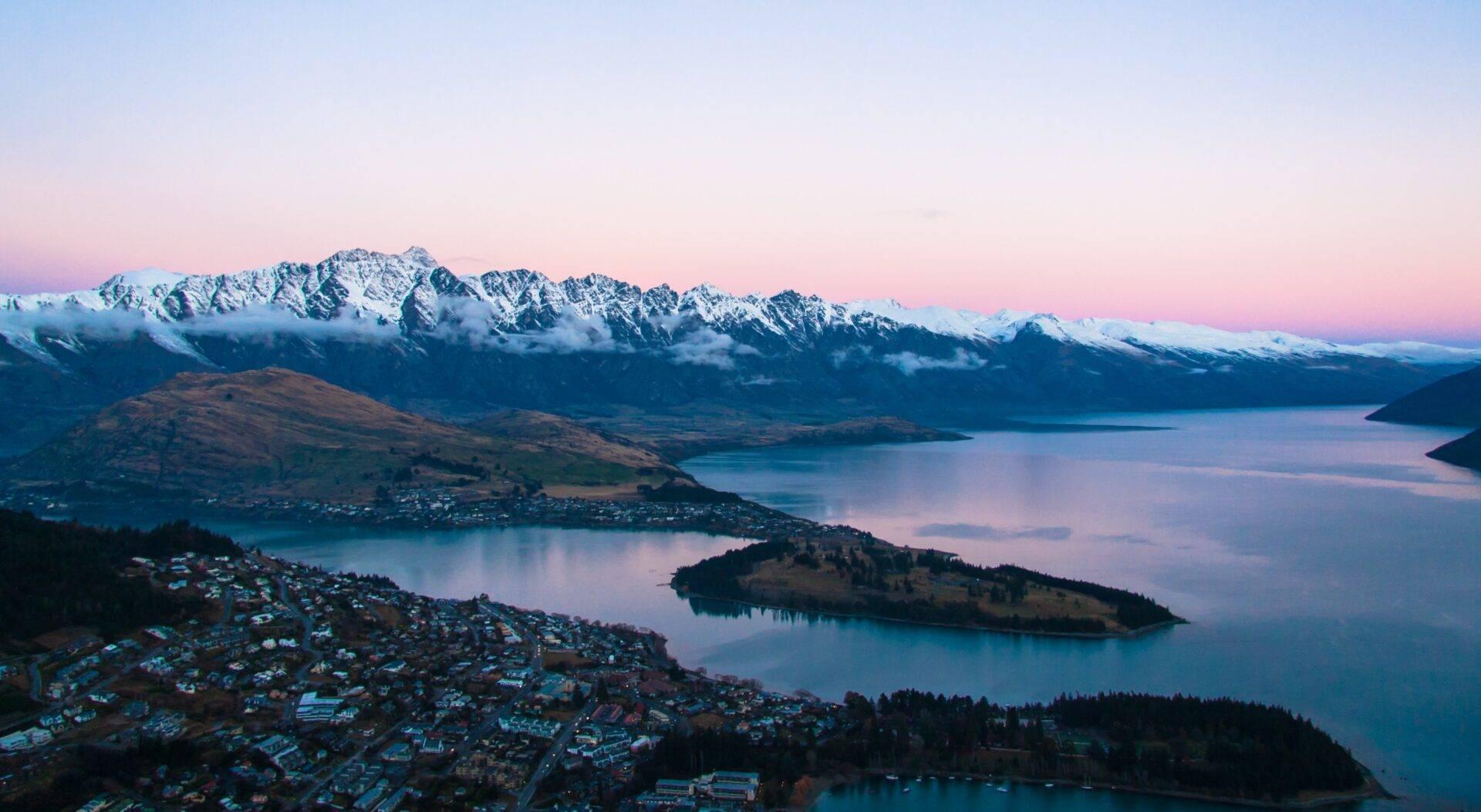 Vue aérienne d’une ville côtière avec lac alpin, montagnes enneigées et péninsules boisées au crépuscule