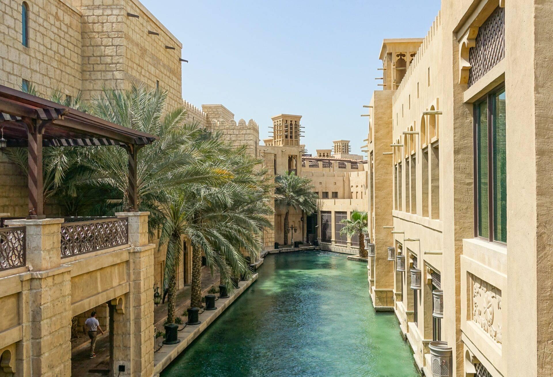 Un canal d’eau turquoise bordé d’immeubles en pierre ocre avec palmiers et architecture traditionnelle du Moyen-Orient
