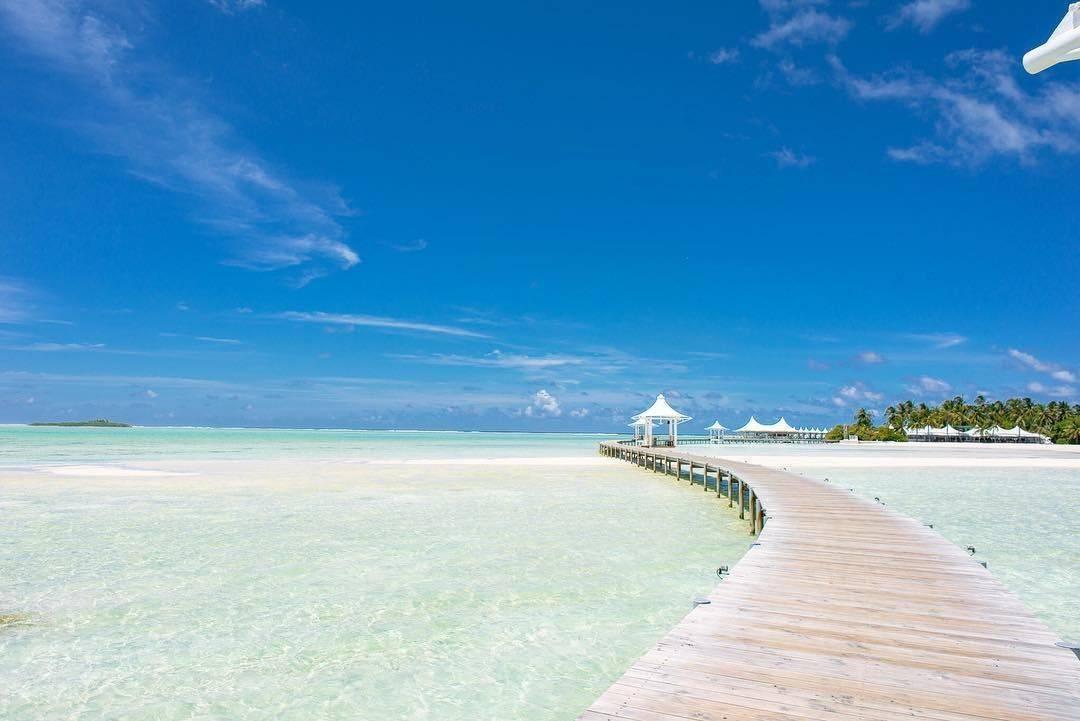 Une jetée en bois s’étend vers un pavillon blanc sur une plage de sable fin aux eaux turquoise cristallines