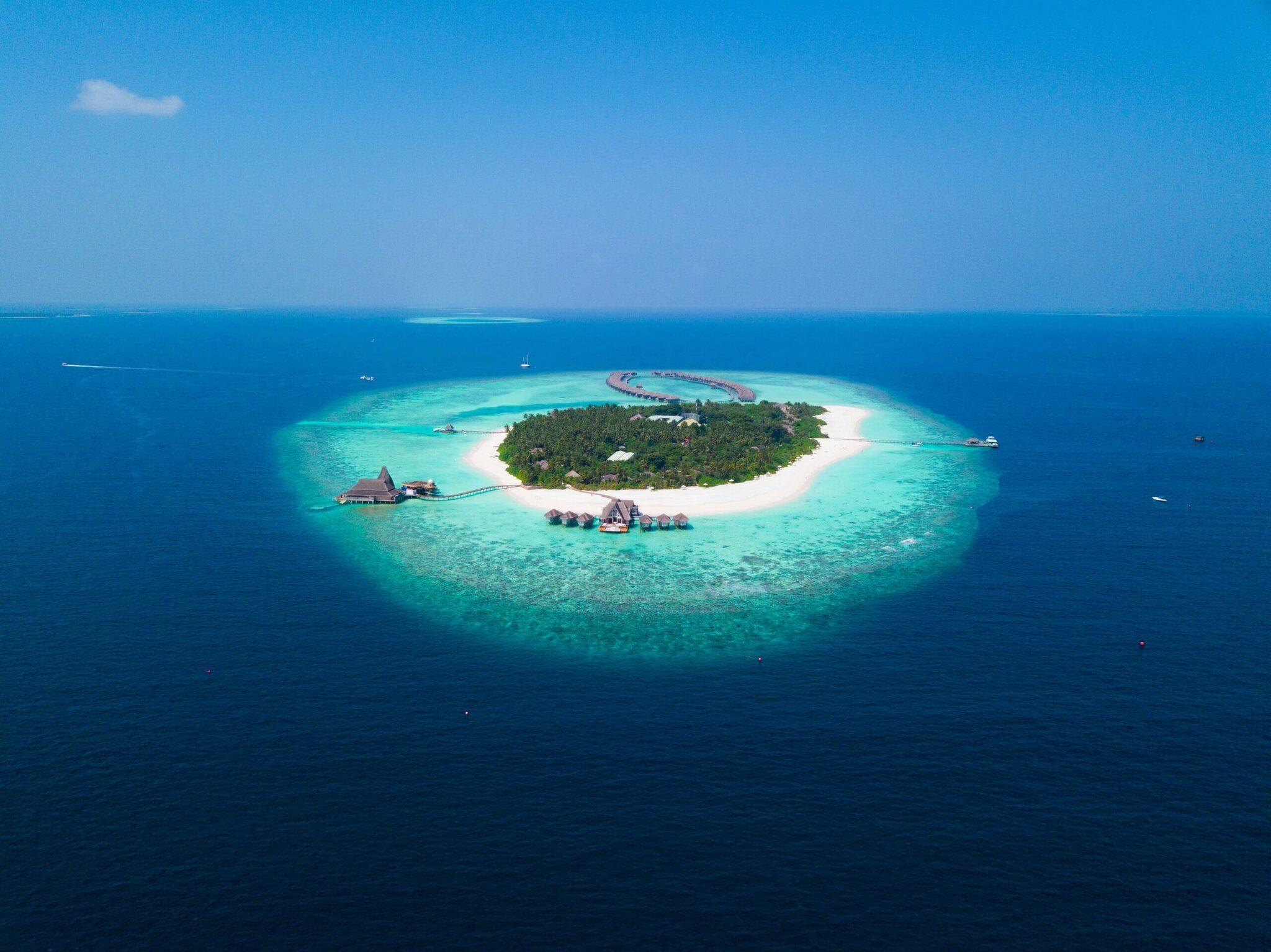 Vue aérienne d’une île tropicale circulaire avec plage blanche, lagon turquoise et bungalows sur pilotis