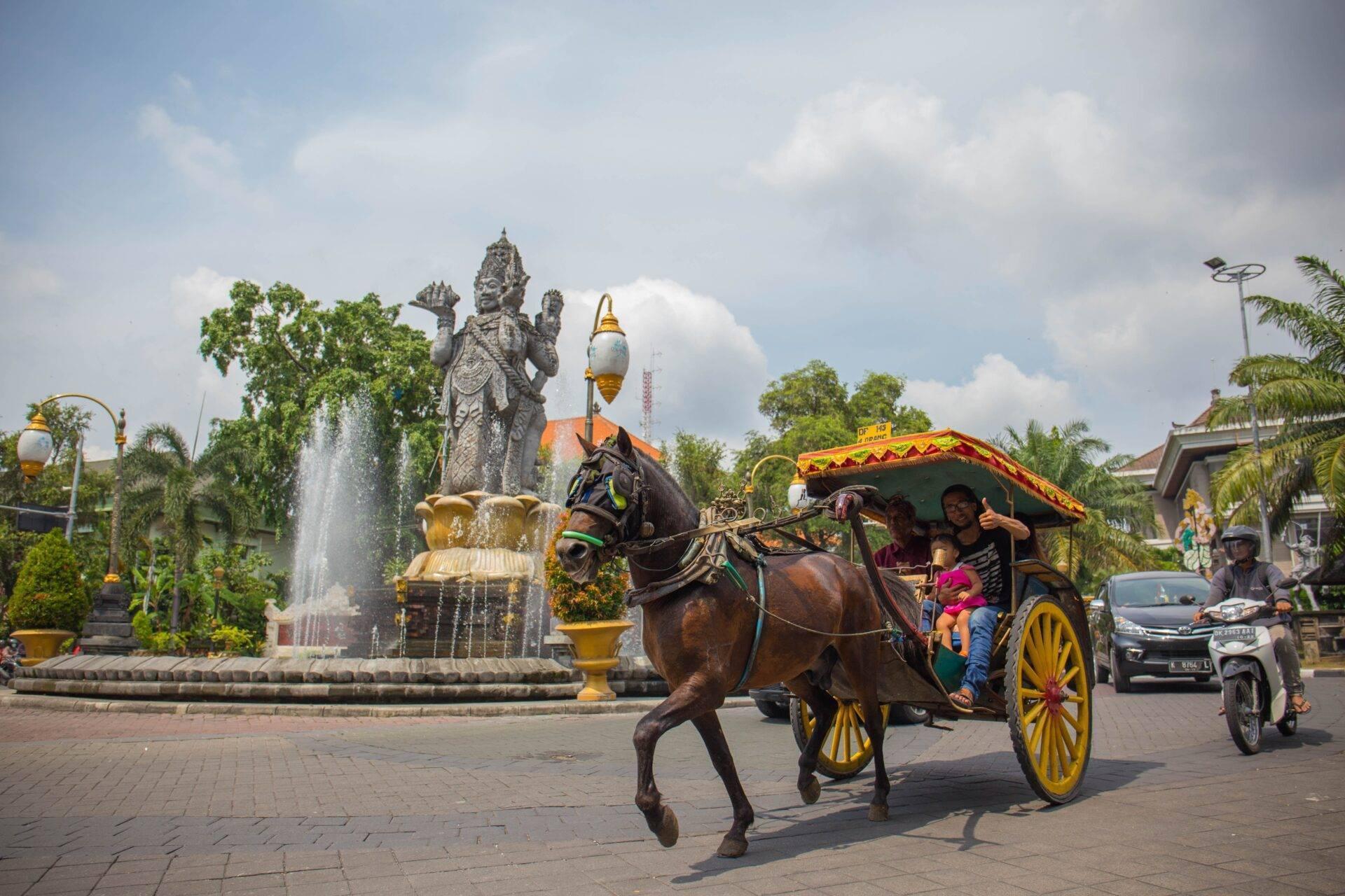 Un char à cheval traditionnel jaune et rouge passe devant une statue divine dans une place publique indonésienne