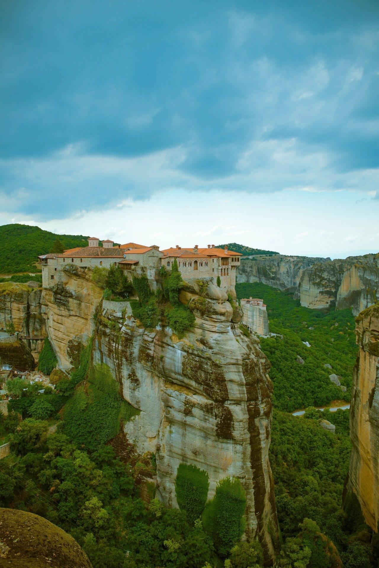 Un monastère perché sur une falaise rocheuse avec toits orangés dominant une vallée verdoyante sous ciel nuageux