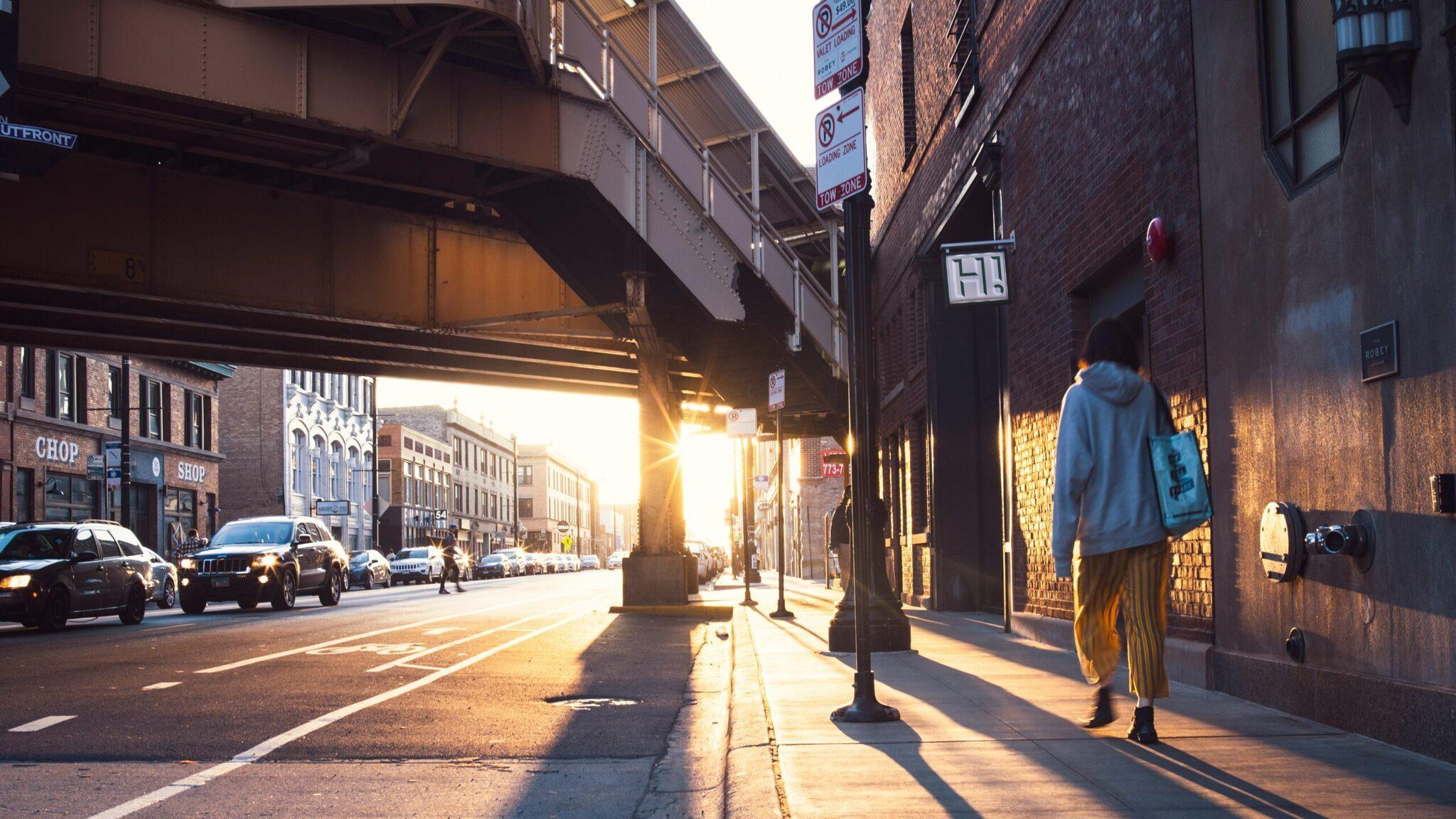 Une rue urbaine américaine sous un viaduc ferroviaire au coucher de soleil, avec piétons et voitures circulant entre bâtiments en brique