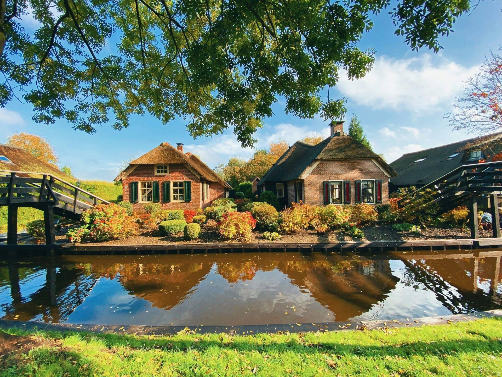 Deux maisons traditionnelles en brique avec toit de chaume se reflètent dans un canal, entourées d’arbres et de végétation automnale